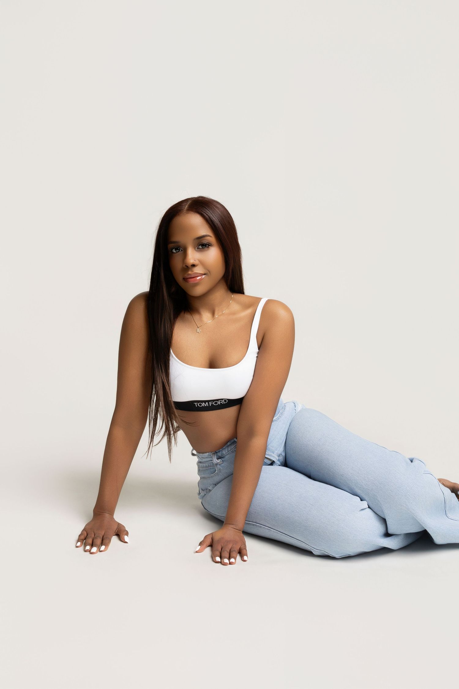 Young woman in white Tom Ford bralette and light blue jeans posing on floor against white background.