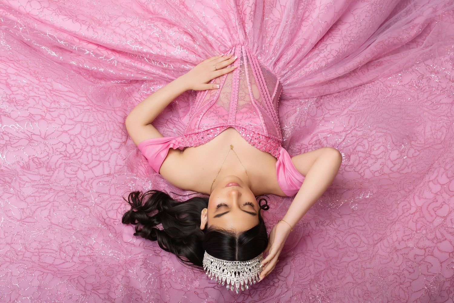 Woman in pink lace dress lying on pink fabric backdrop with elegant pose and dark flowing hair.