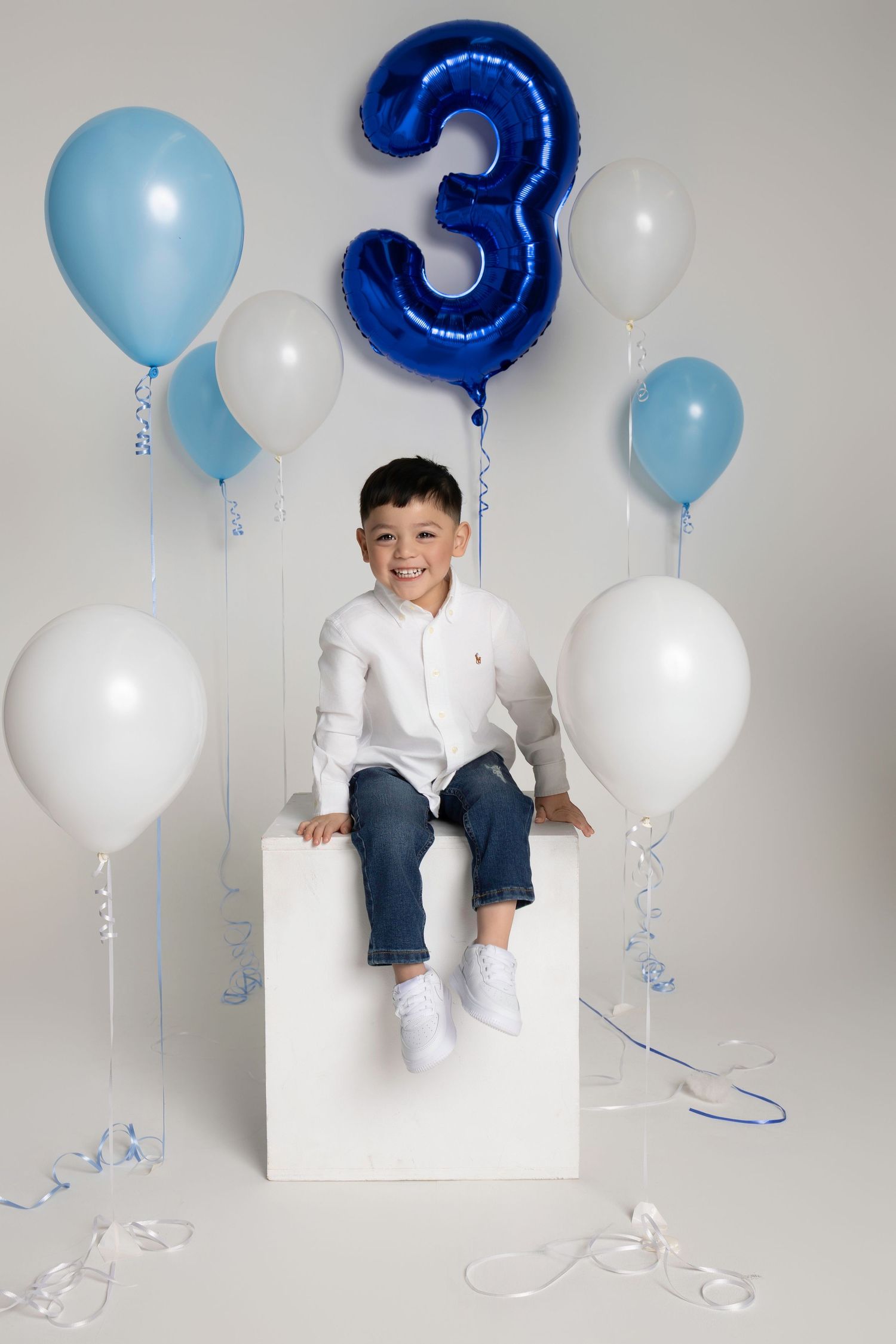 Smiling boy celebrates 3rd birthday sitting on white cube surrounded by blue and white balloons.