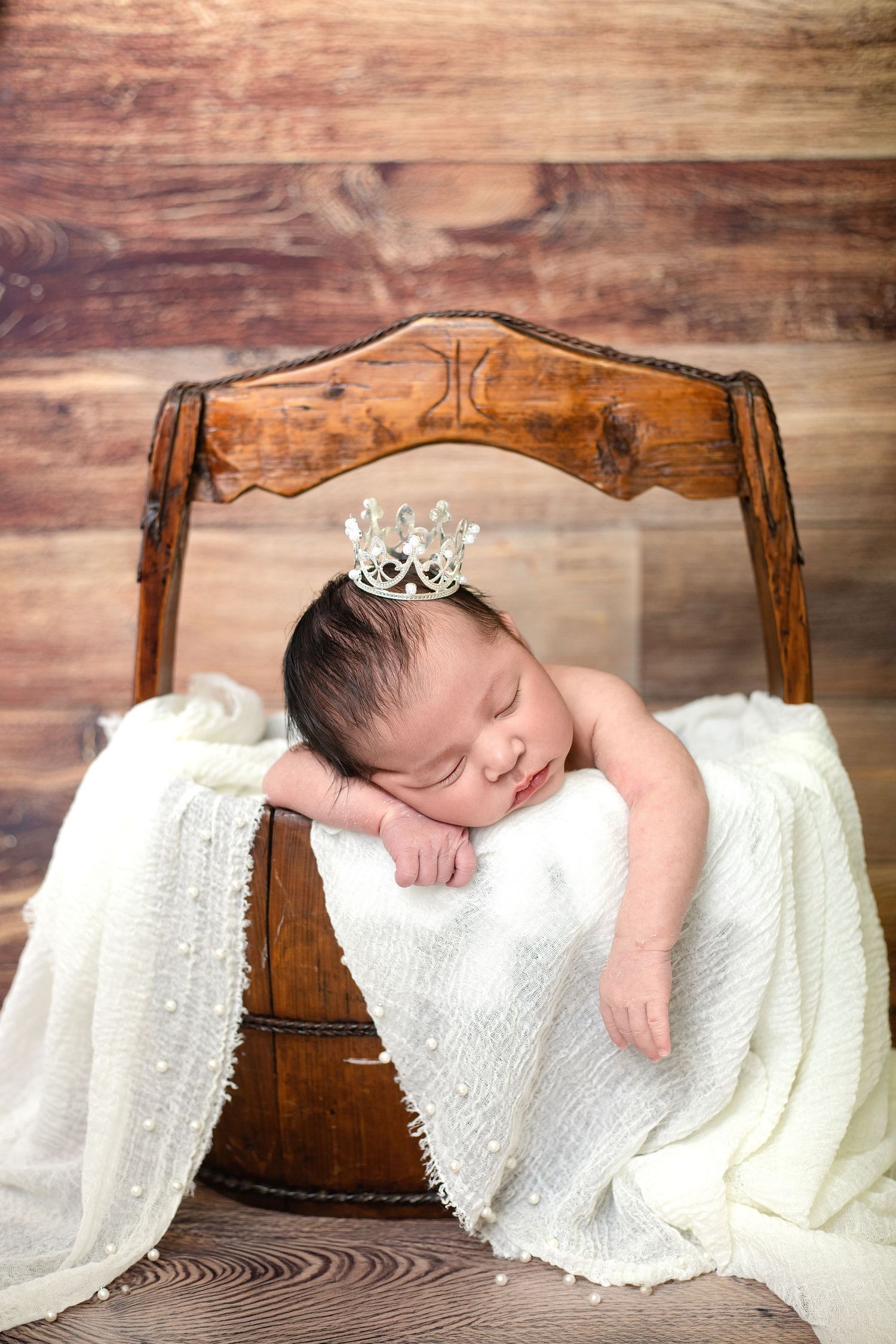 Newborn baby sleeping peacefully in a wooden bucket, wearing a tiny crown, wrapped in a white lace blanket.