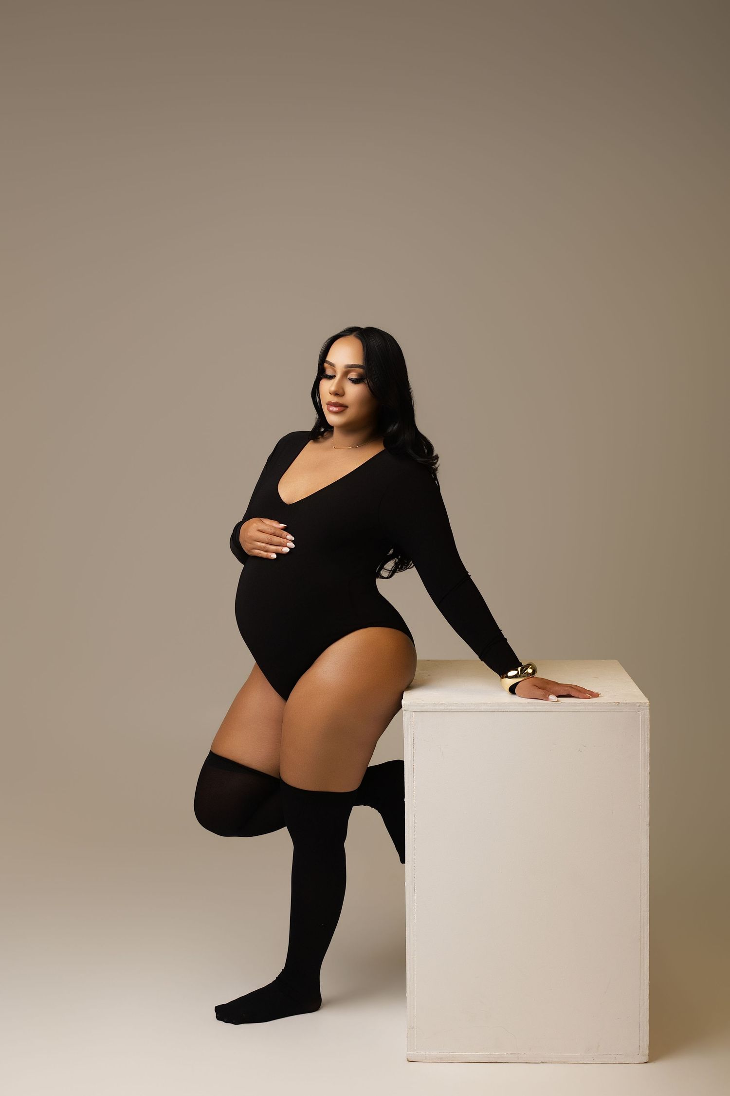 Pregnant woman in black bodysuit and knee-high socks poses elegantly leaning on white cube in studio.