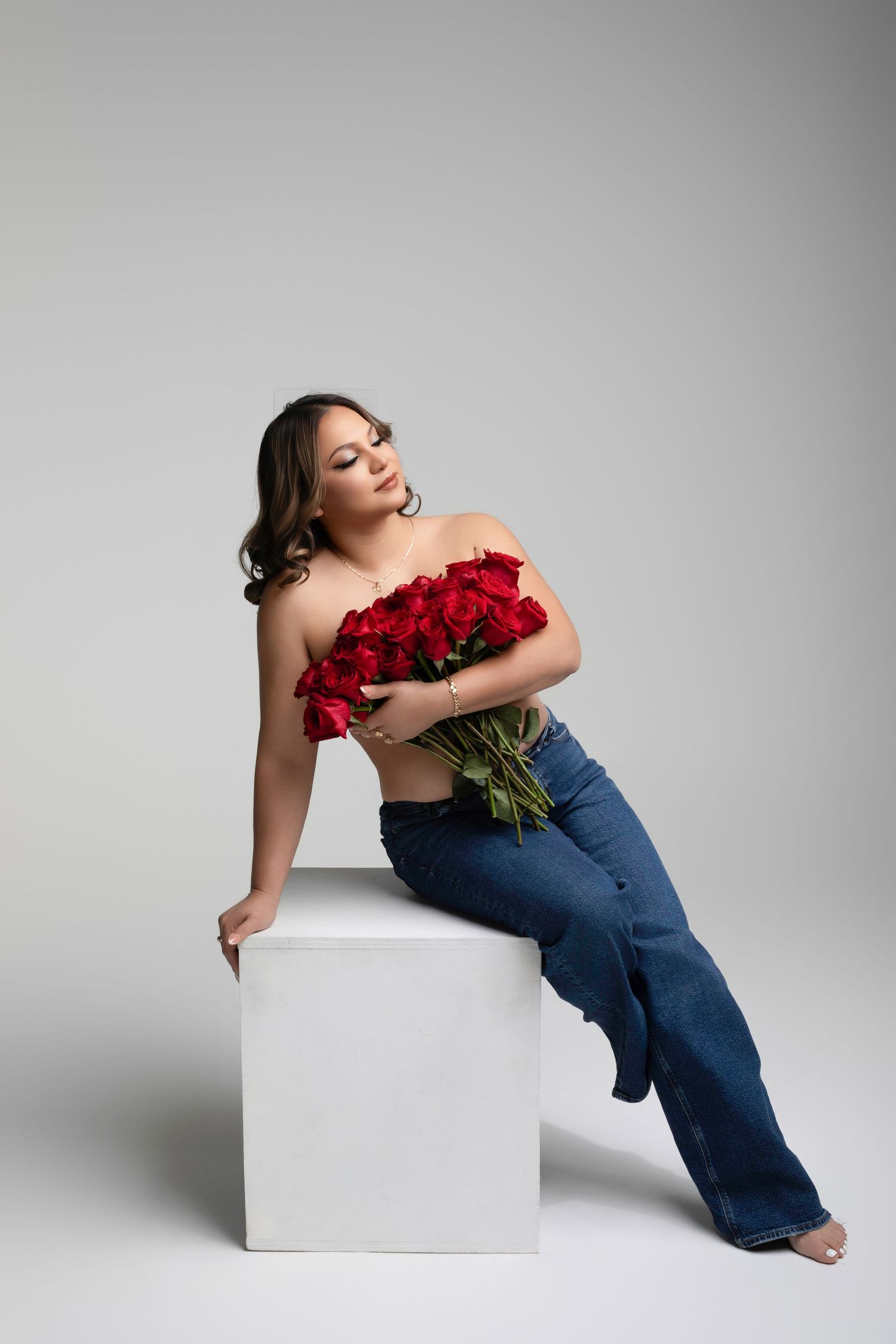 Woman in jeans holding red roses bouquet, sitting on white cube in studio, elegant pose with bare shoulders.