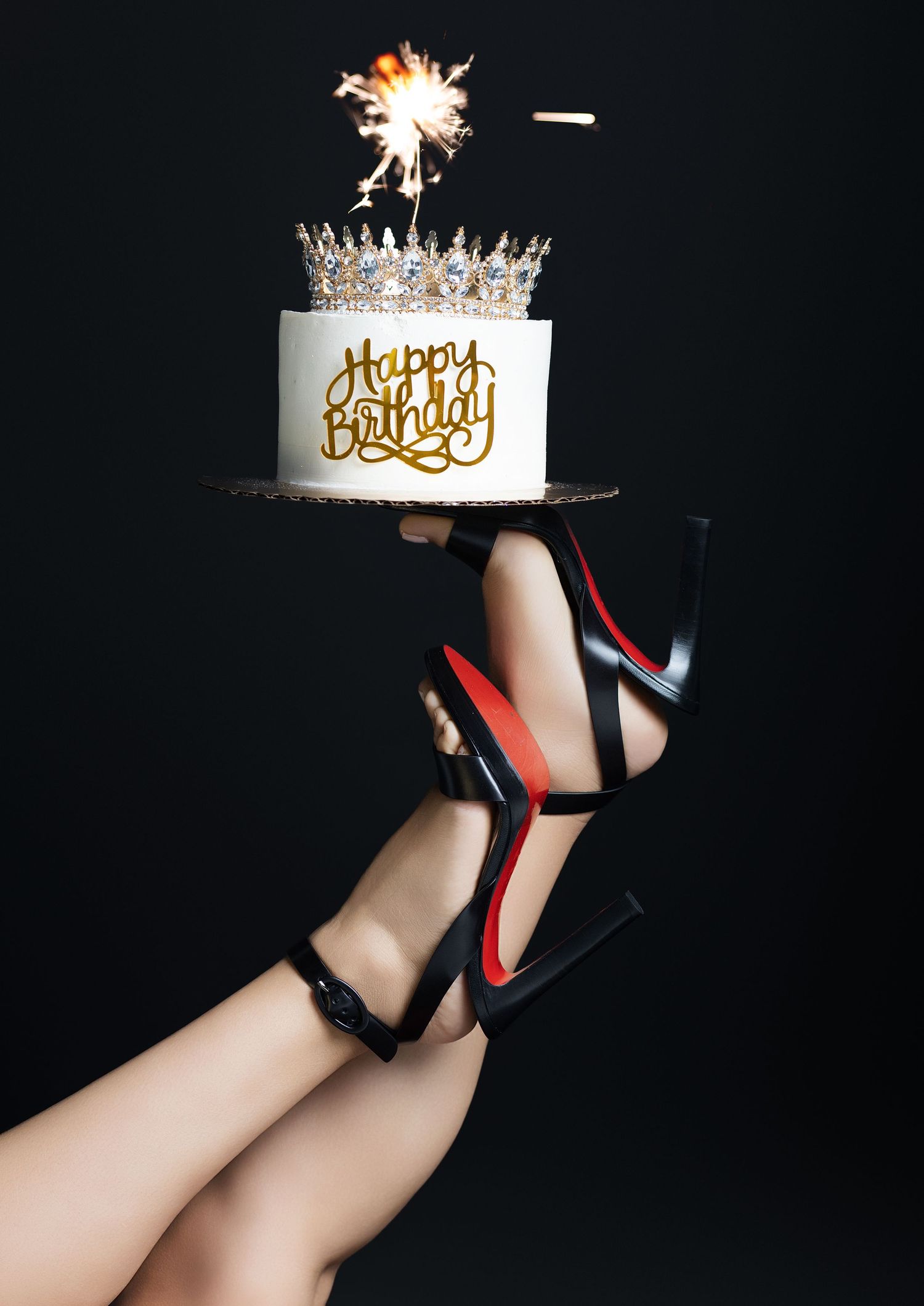 Legs in black high heels balancing a white Happy Birthday cake with crown topper and sparkler on black background.
