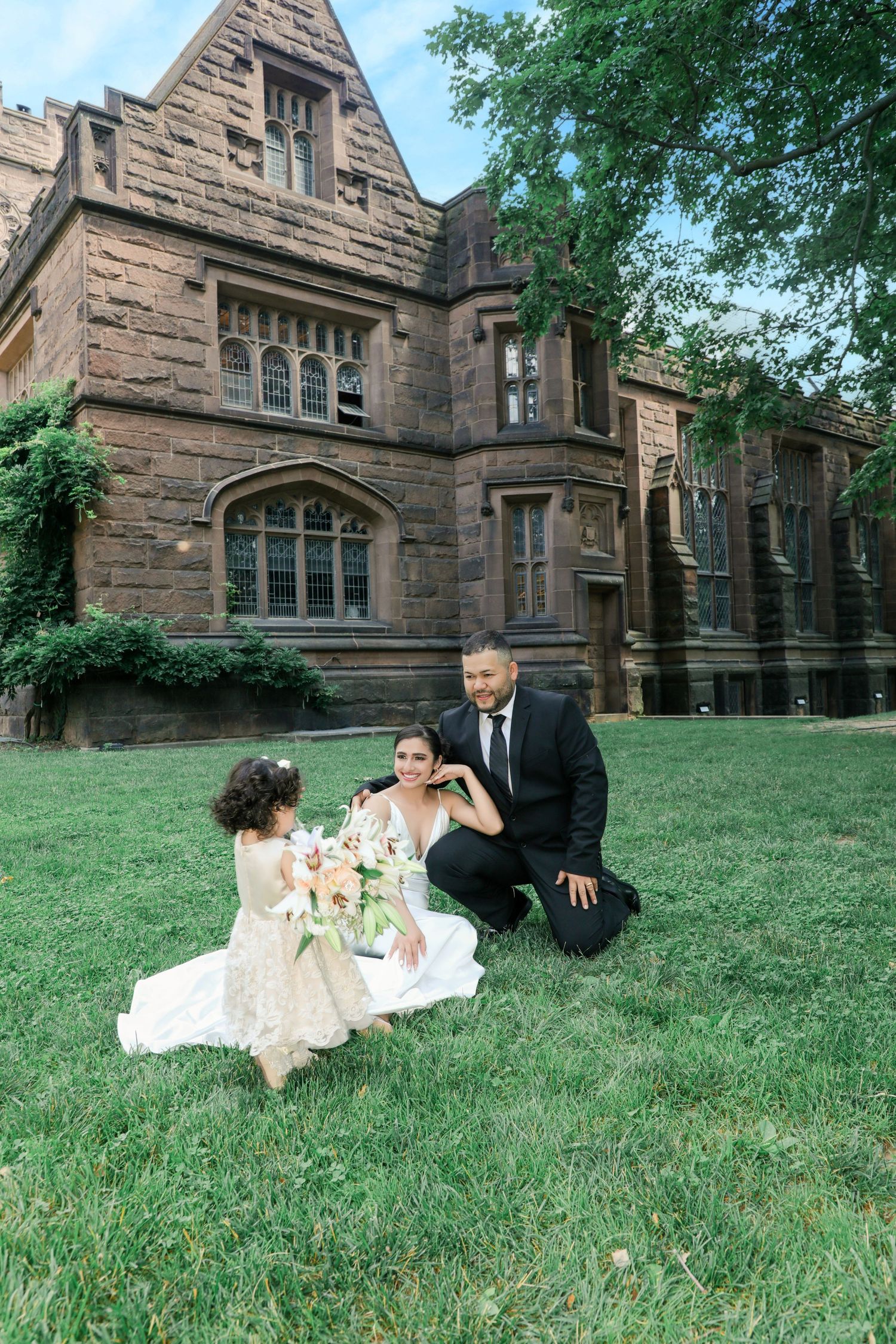 Bride and groom kneeling on grass to greet a flower girl outside a historic stone mansion.