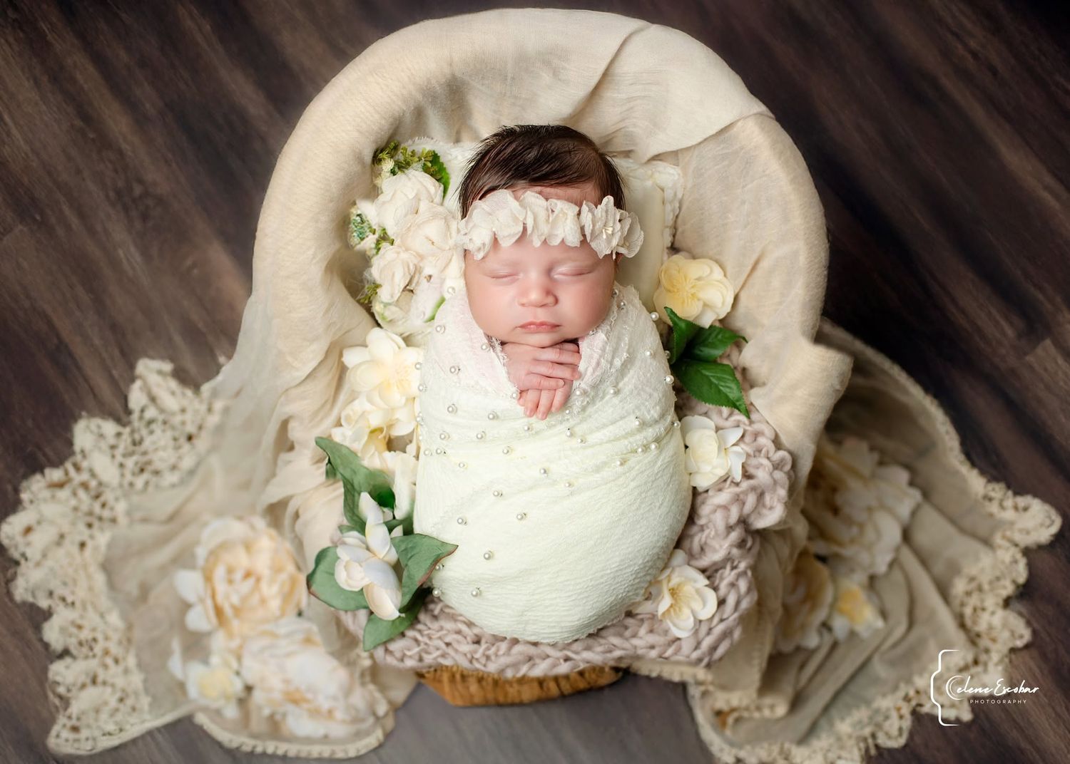 Newborn baby girl swaddled in cream wrap, sleeping in a basket with white floral headband and flower decorations.