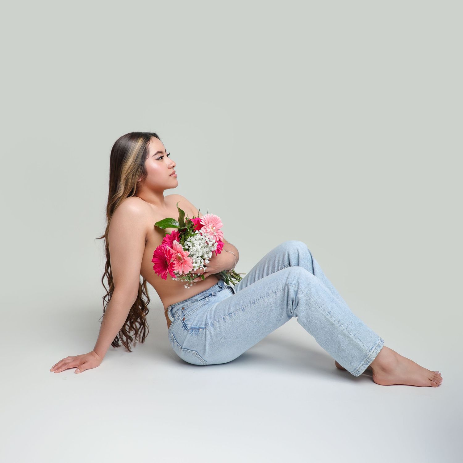 Young woman sitting on floor holding pink and white flower bouquet, wearing light blue jeans against gray background.