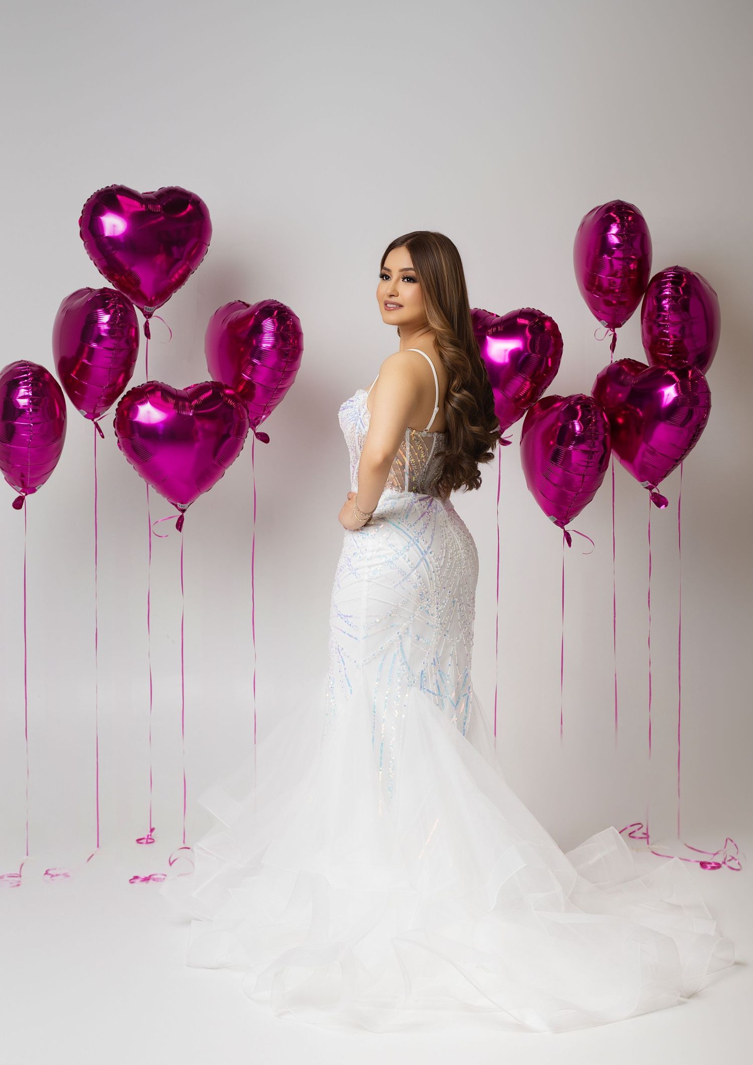Woman in white mermaid gown poses elegantly surrounded by pink heart-shaped foil balloons on white background.