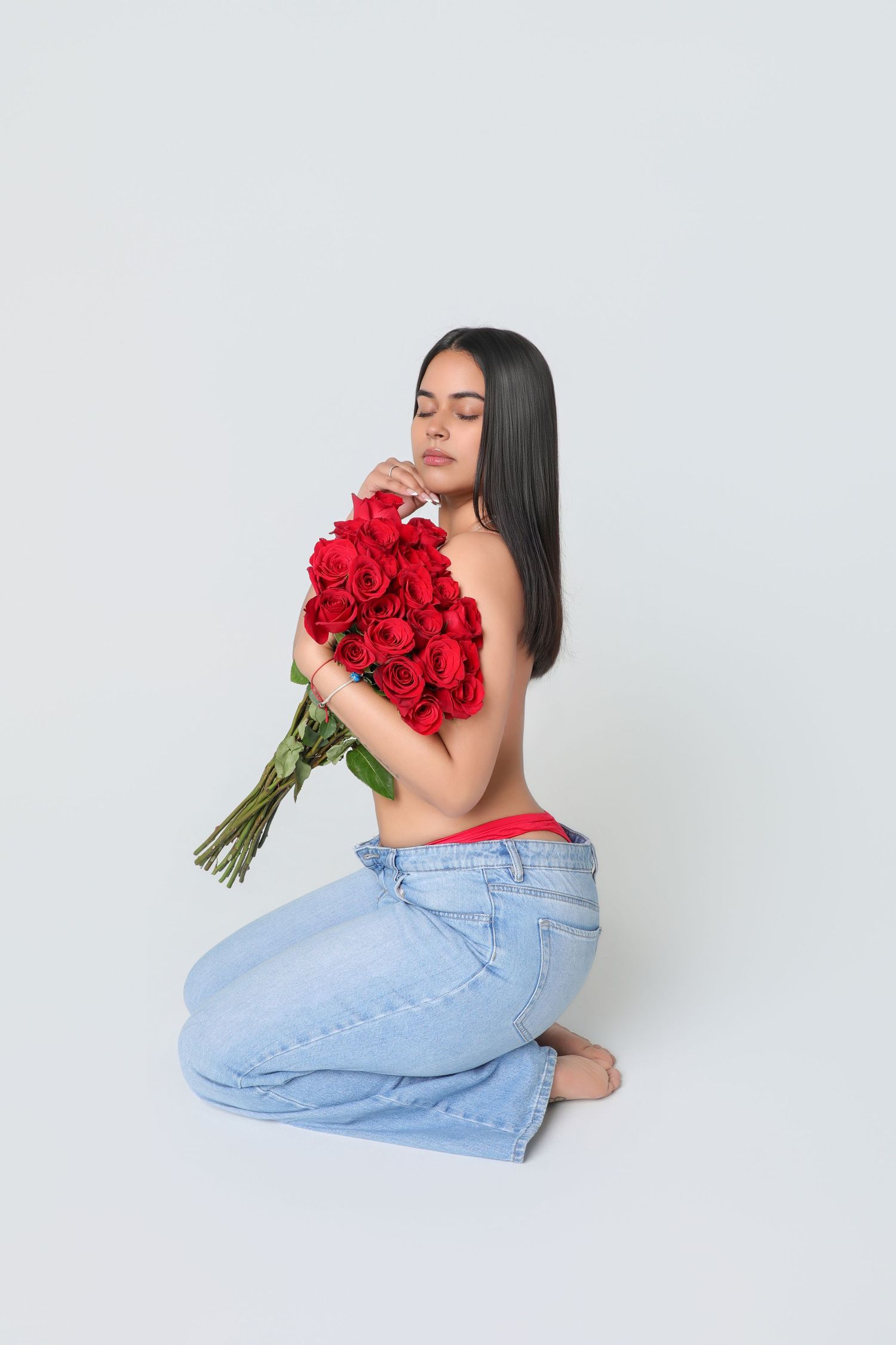 Woman kneeling with bouquet of red roses, wearing jeans, eyes closed, white background.