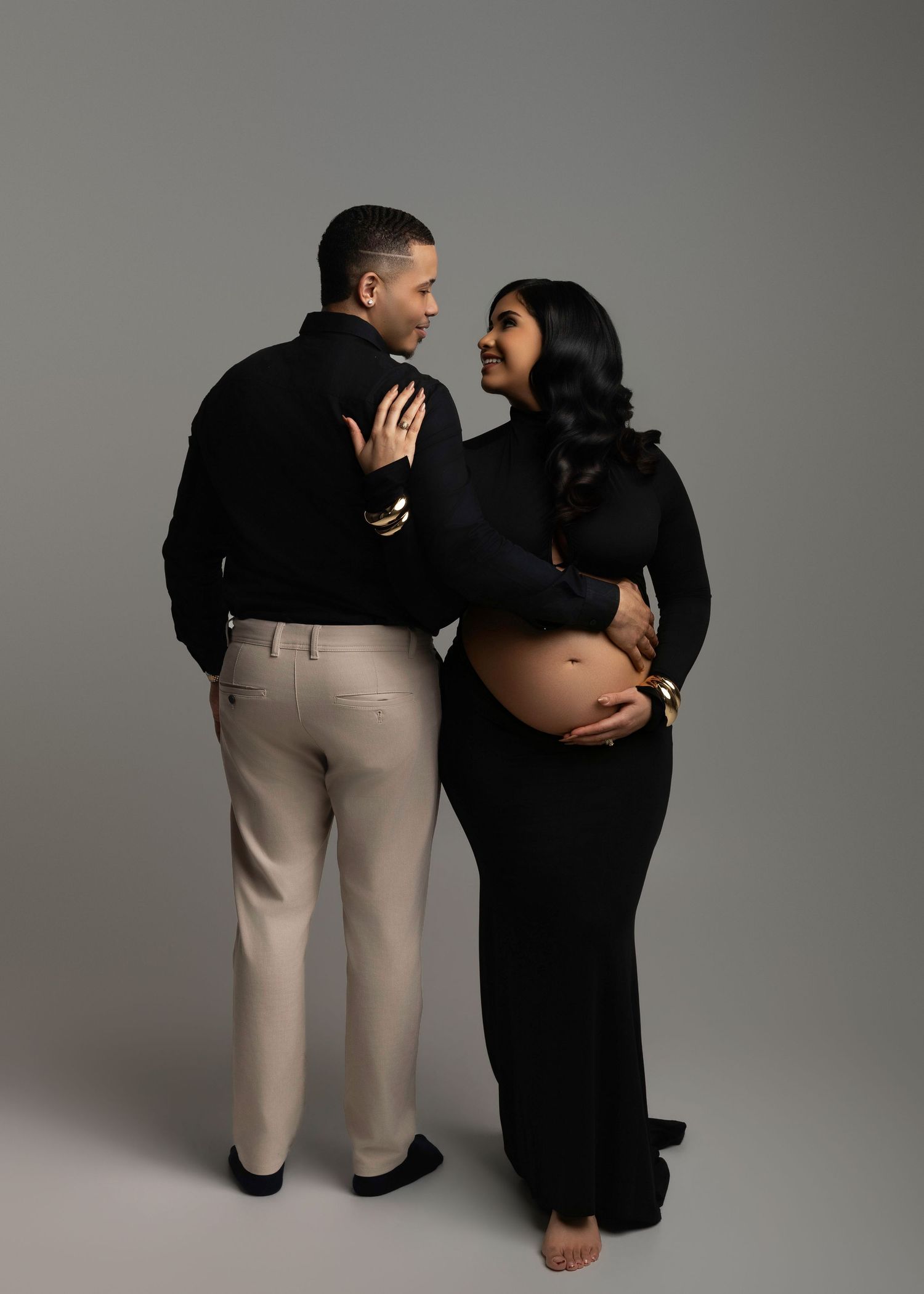 Couple in black outfits sharing a tender moment, pregnant woman cradling baby bump in a studio maternity photo shoot.