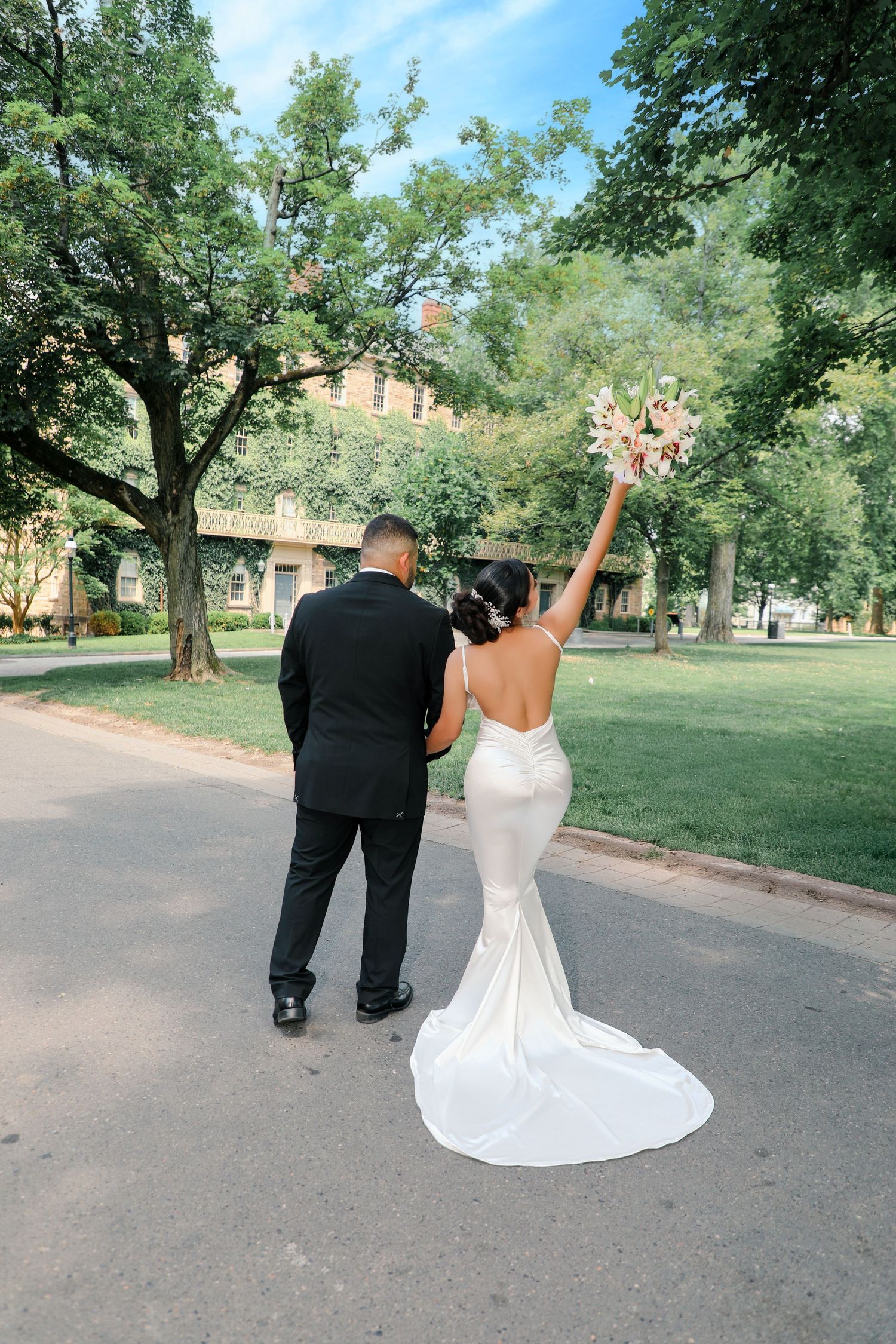Bride raises bouquet joyfully while walking hand-in-hand with groom along tree-lined path.