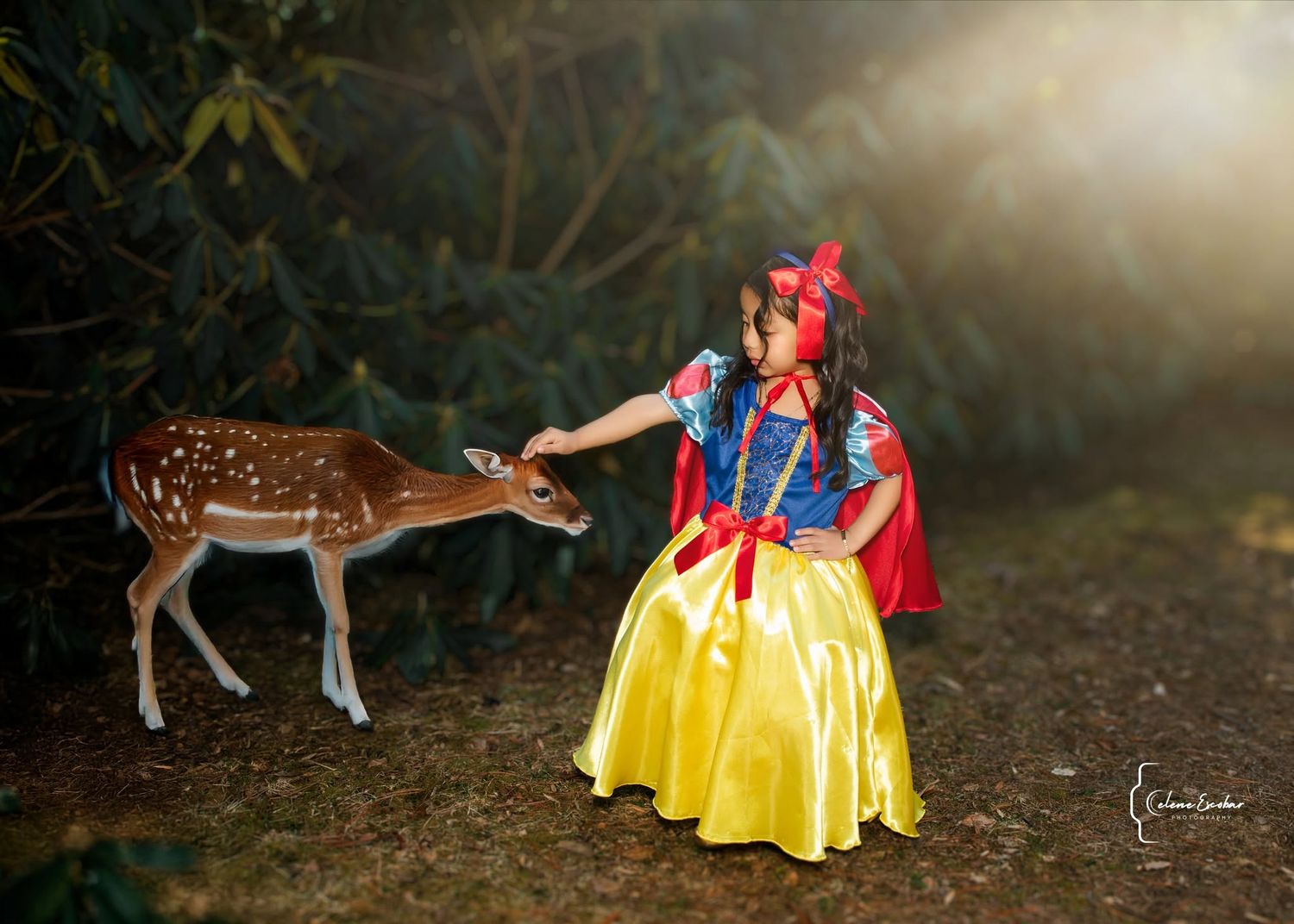 A little girl in a Snow White costume gently feeds a spotted fawn in an enchanted forest setting.
