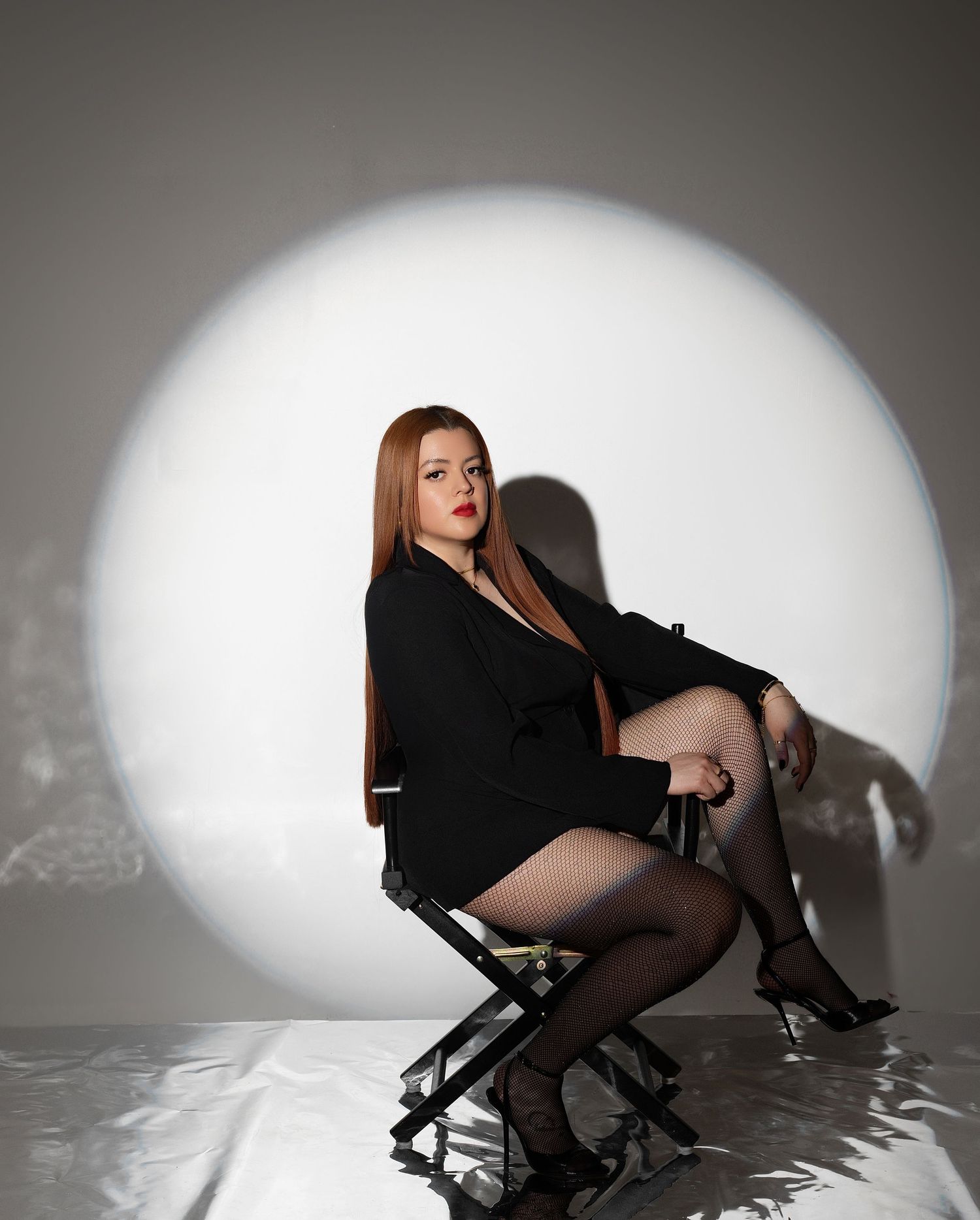Woman in black blazer dress and heels sits on director's chair before circular spotlight backdrop.