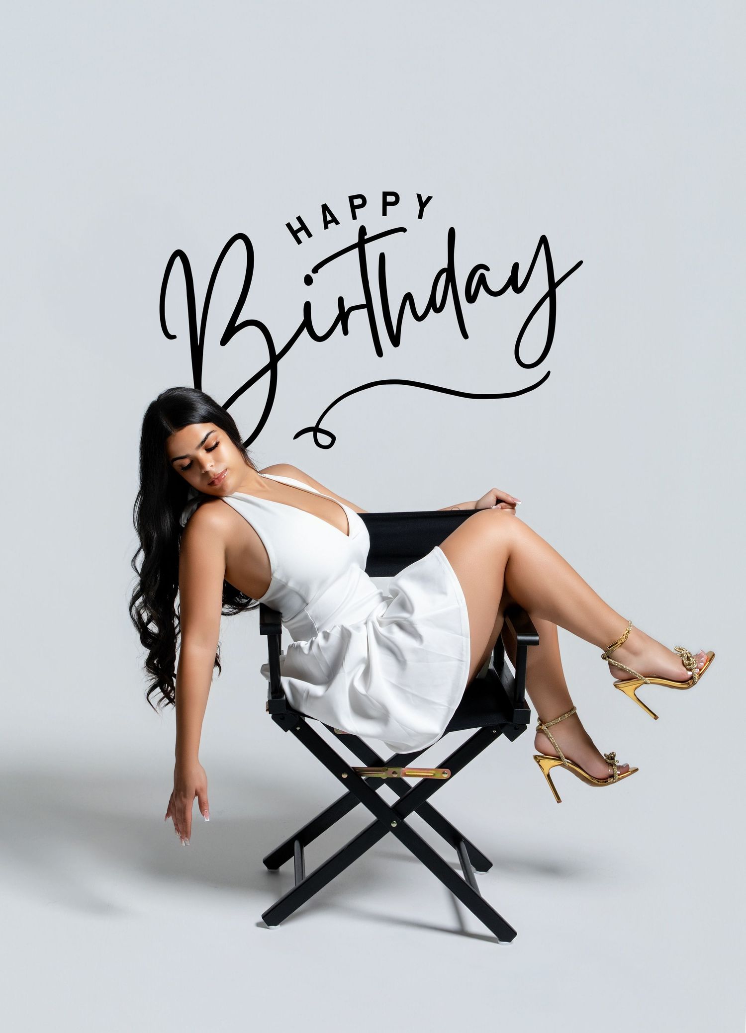 Woman in white dress seated on director's chair with Happy Birthday text overlay on light gray background.