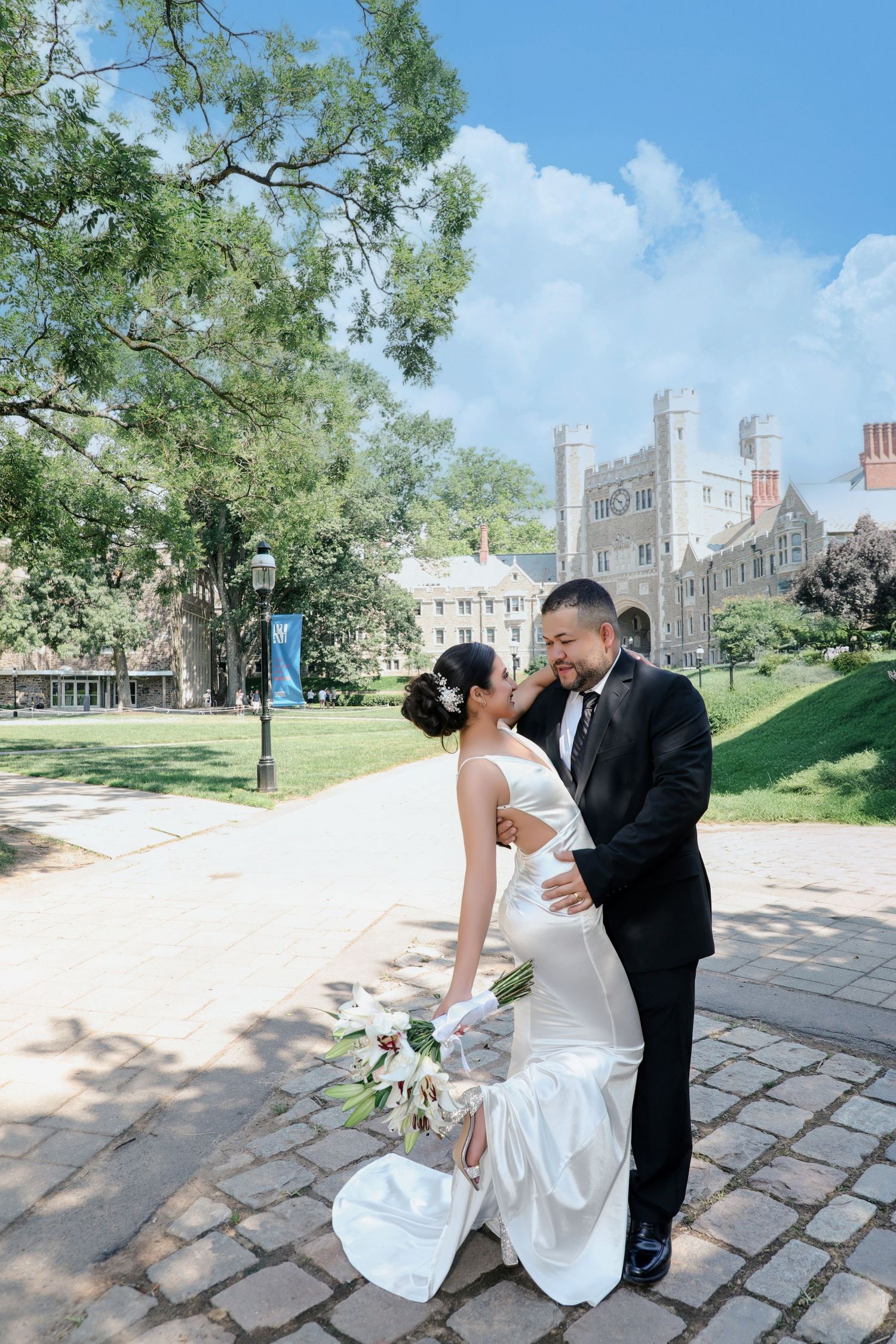 Newlywed couple sharing a romantic moment on a sunny campus pathway with castle architecture.