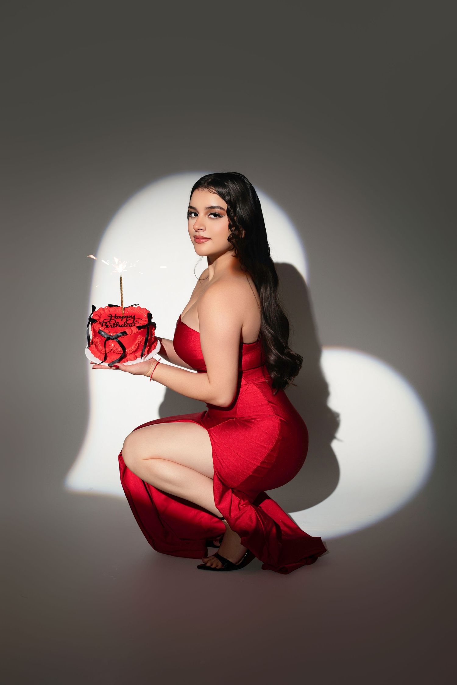 Woman in red strapless dress crouching, holding heart-shaped rose cake with sparkler, against heart-shaped light backdrop.
