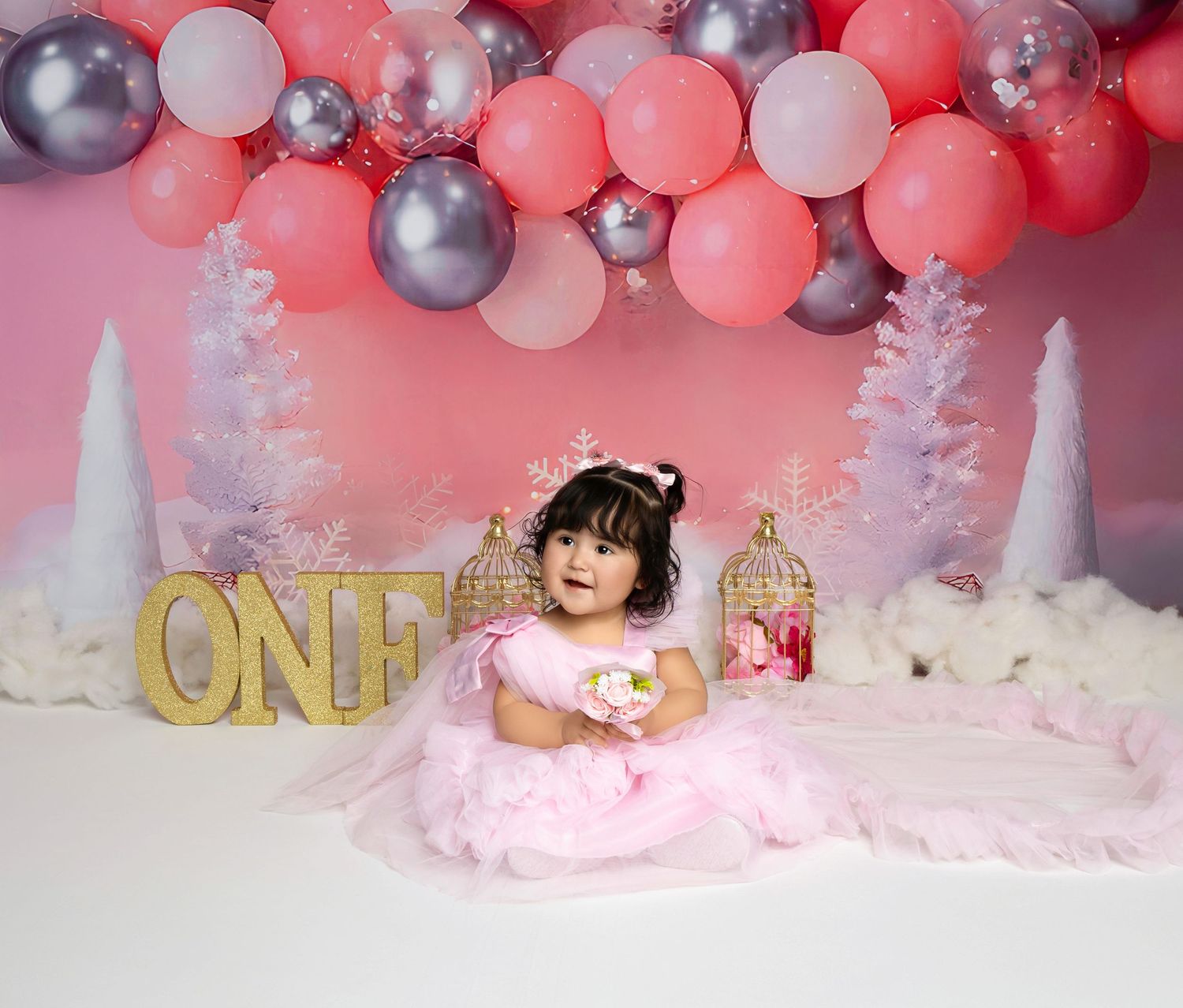 A baby girl in a pink tutu dress sits before a pink balloon arch and ONE sign at her first birthday cake smash.