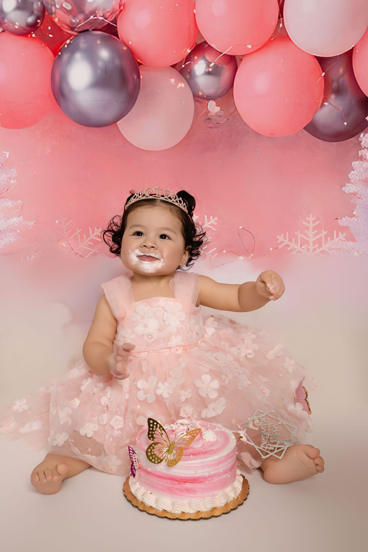 Baby girl in pink dress celebrates first birthday with butterfly cake and pink balloon garland backdrop.