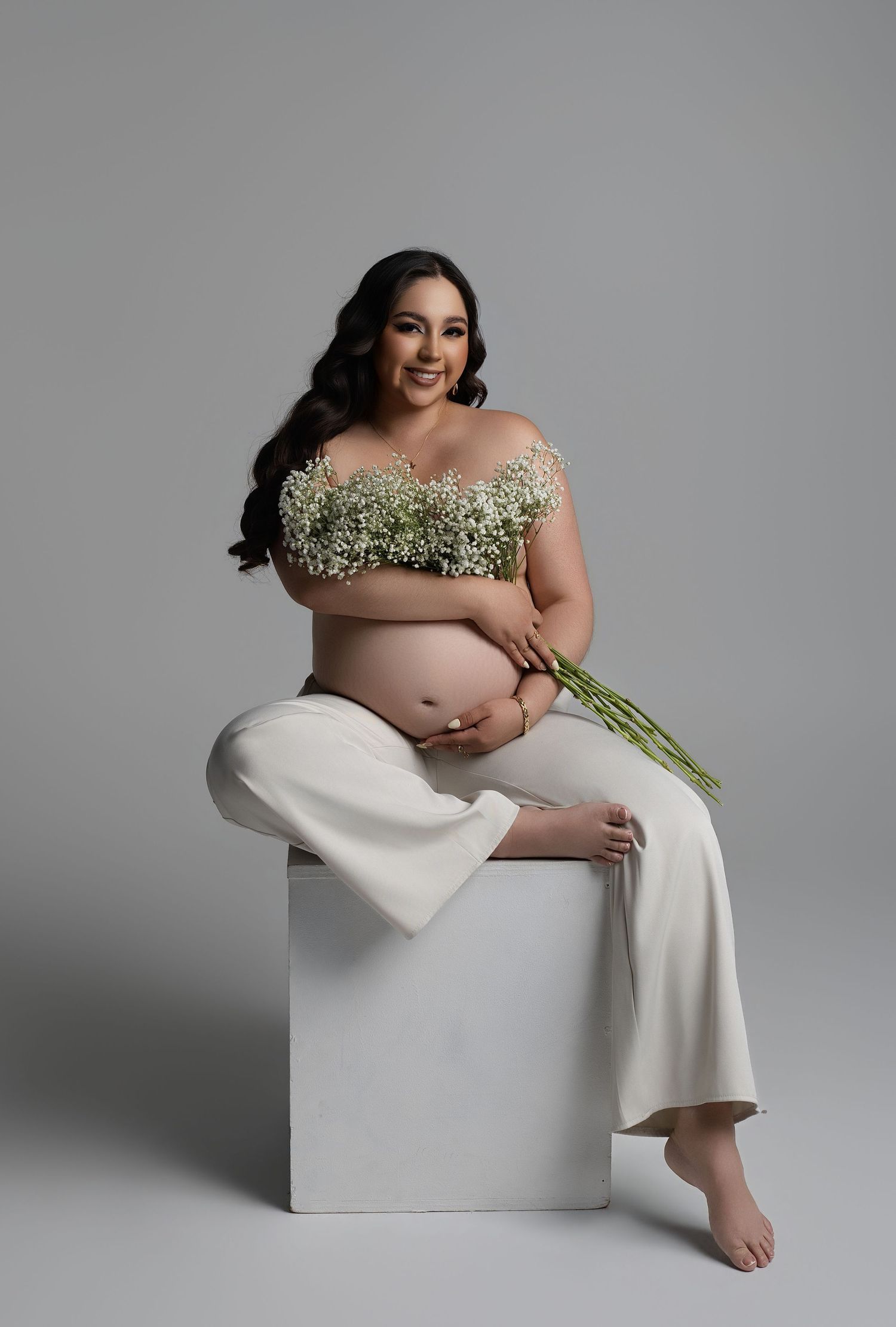 Pregnant woman in white pants holding baby's breath flowers, seated on white cube for maternity photo shoot.