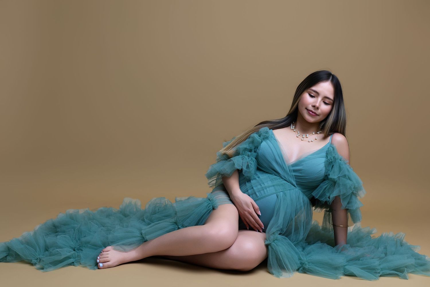Pregnant woman in teal tulle maternity gown posing gracefully on floor against warm beige background.