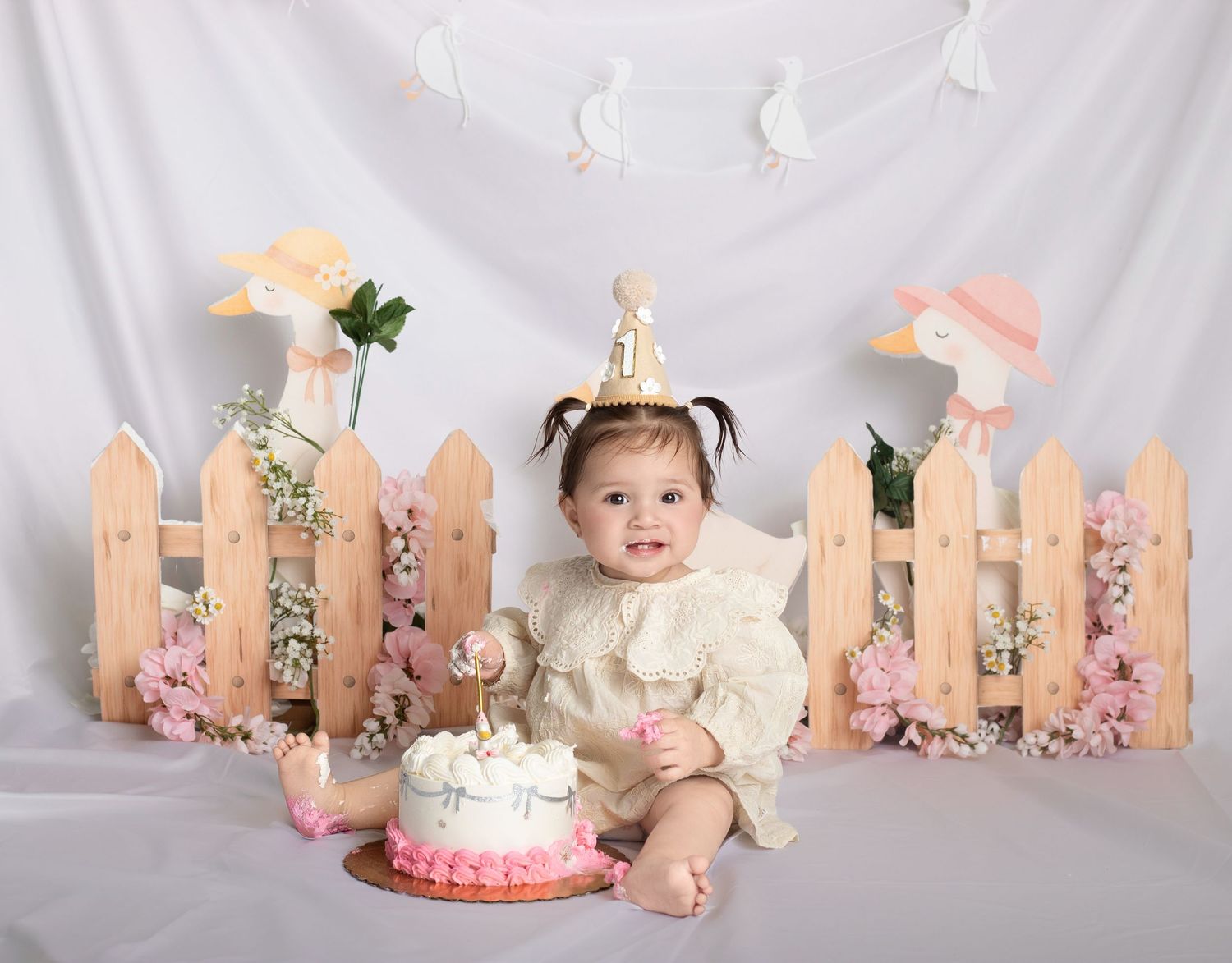 Baby girl in birthday hat smashing pink cake at first birthday photo shoot with floral fence backdrop.