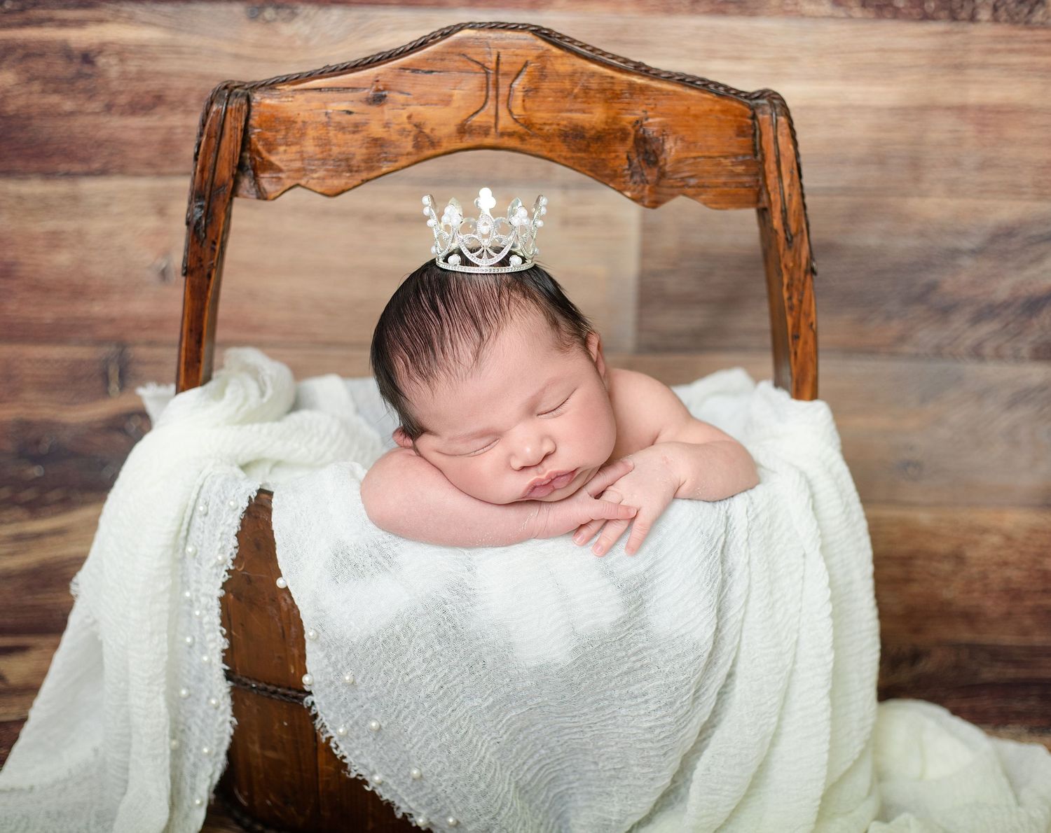Sleeping newborn baby wearing a tiny crystal crown, wrapped in white lace, nestled in a rustic wooden mini bed.