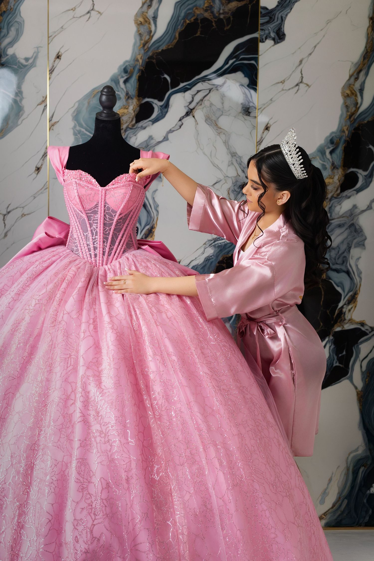 Woman in pink satin robe and tiara adjusting an elegant pink ball gown on a mannequin in a luxury studio.