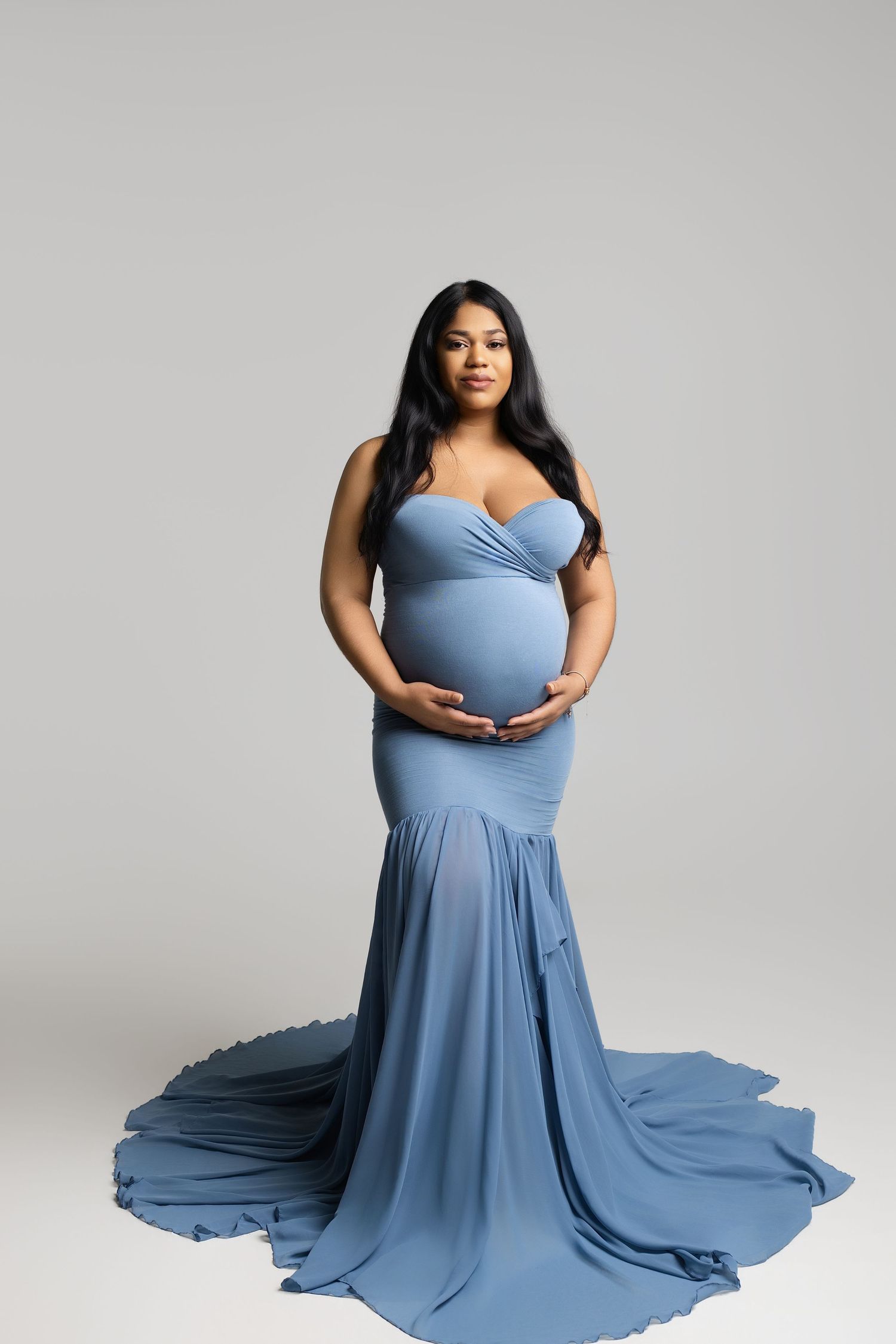 Pregnant woman in elegant blue mermaid maternity gown cradling baby bump, studio portrait photography.