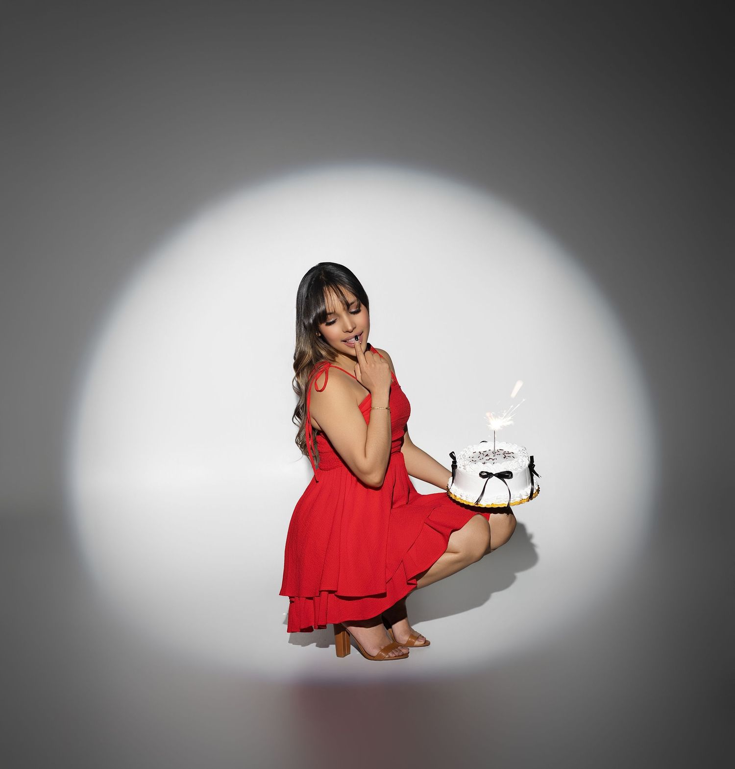 Woman in red dress crouching under spotlight, holding a small decorated birthday cake with candles.