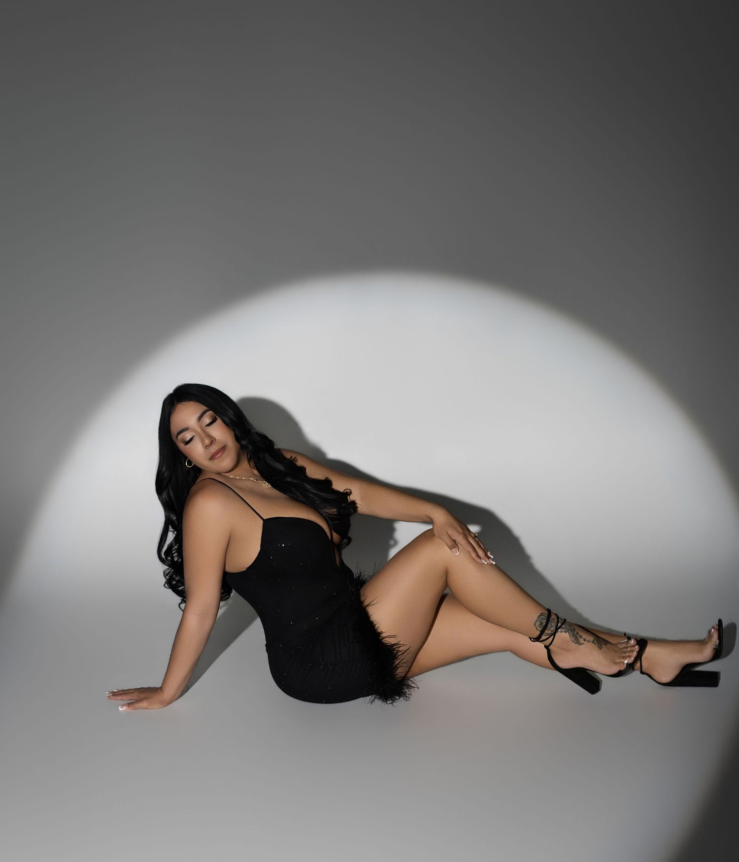 Woman in black mini dress and strappy heels poses on floor under dramatic spotlight in professional studio photo.