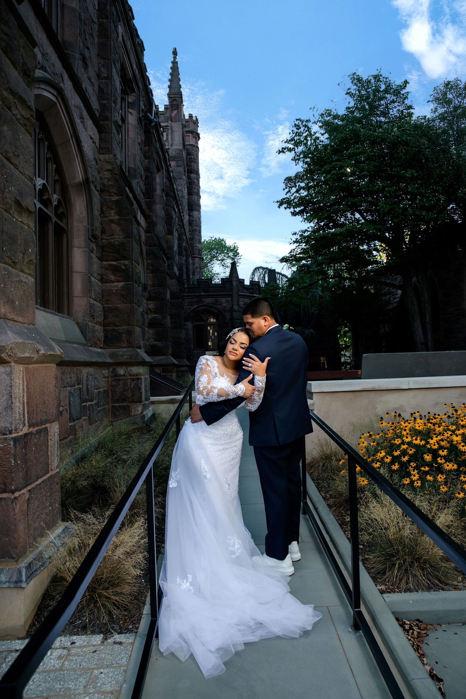 Bride and groom embrace on a bridge beside a Gothic stone church, surrounded by lush greenery and yellow flowers.