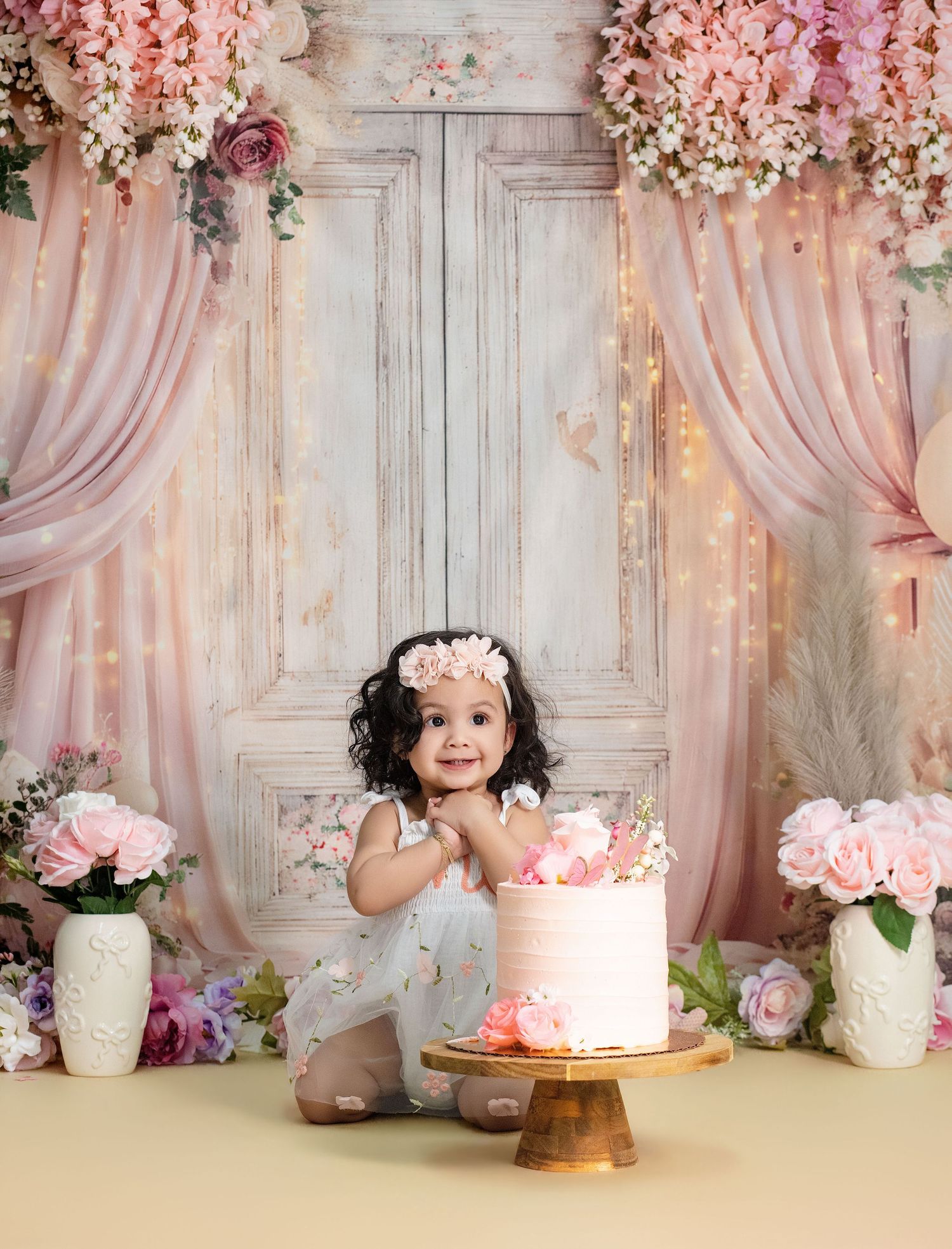 Toddler girl in floral dress smiling beside a pink birthday cake, surrounded by roses and pink curtains.