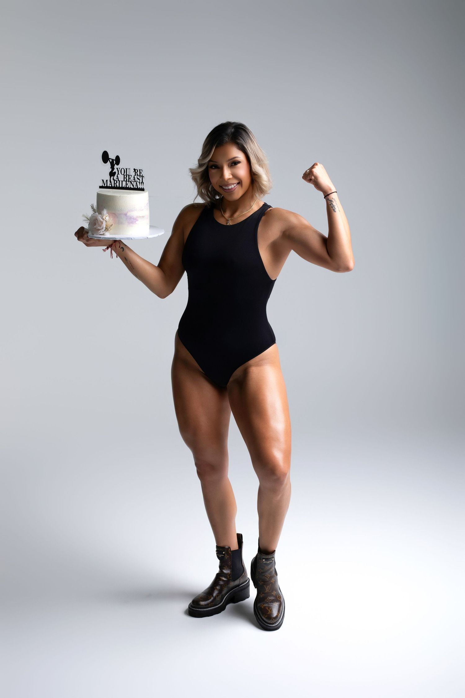 Muscular woman in black bodysuit flexing while holding a decorated birthday cake, wearing brown boots on gray background.