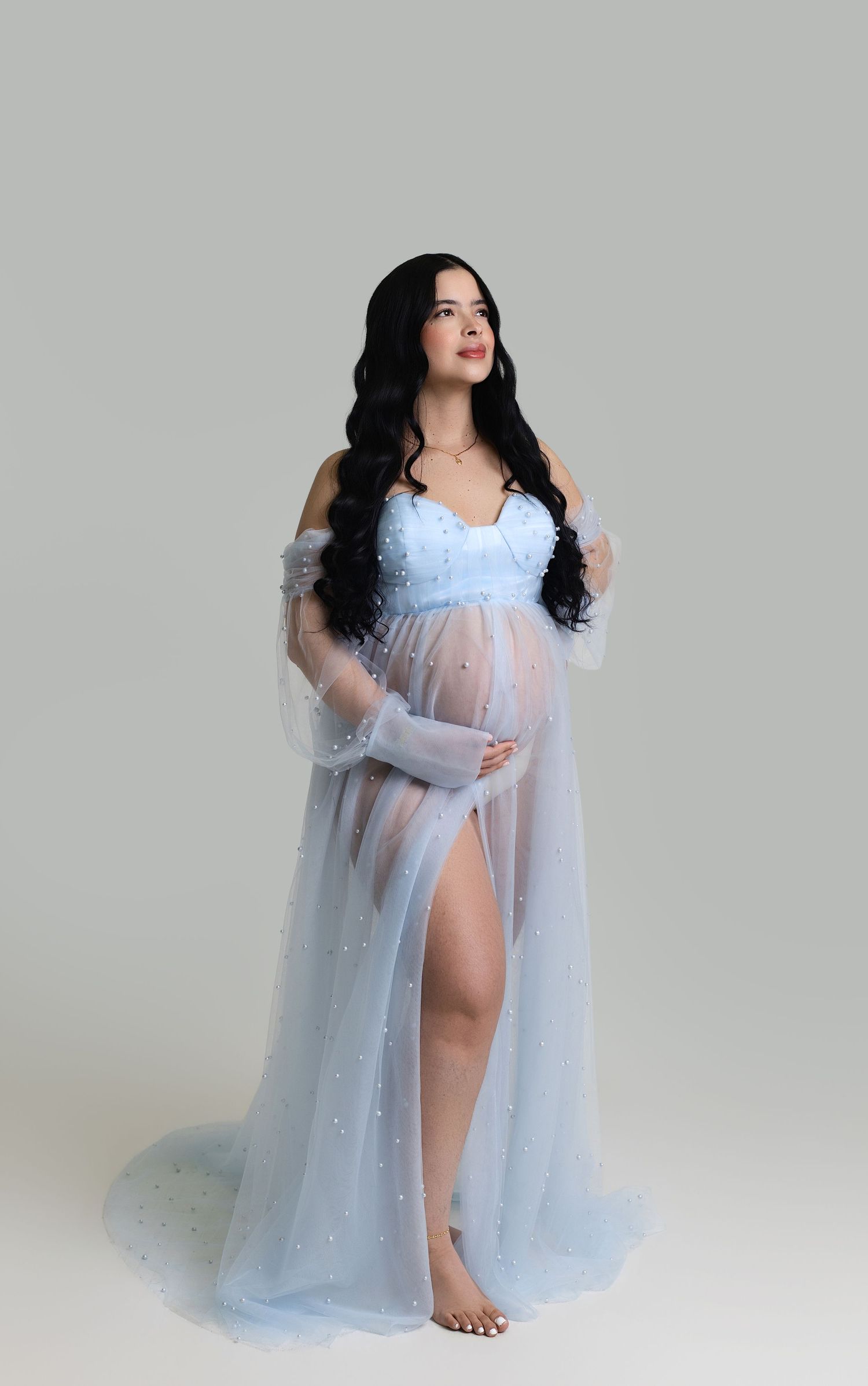 Pregnant woman in sheer light blue maternity gown with pearl details, posing gracefully in a studio setting.
