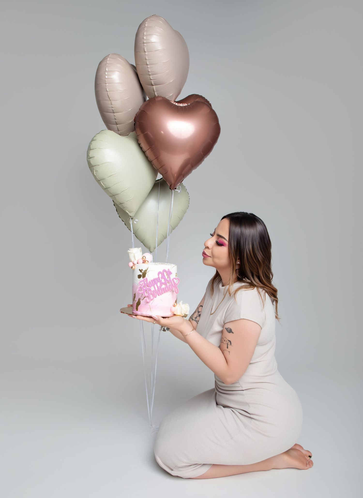 A woman kneels holding a birthday cake with heart-shaped balloons overhead.
