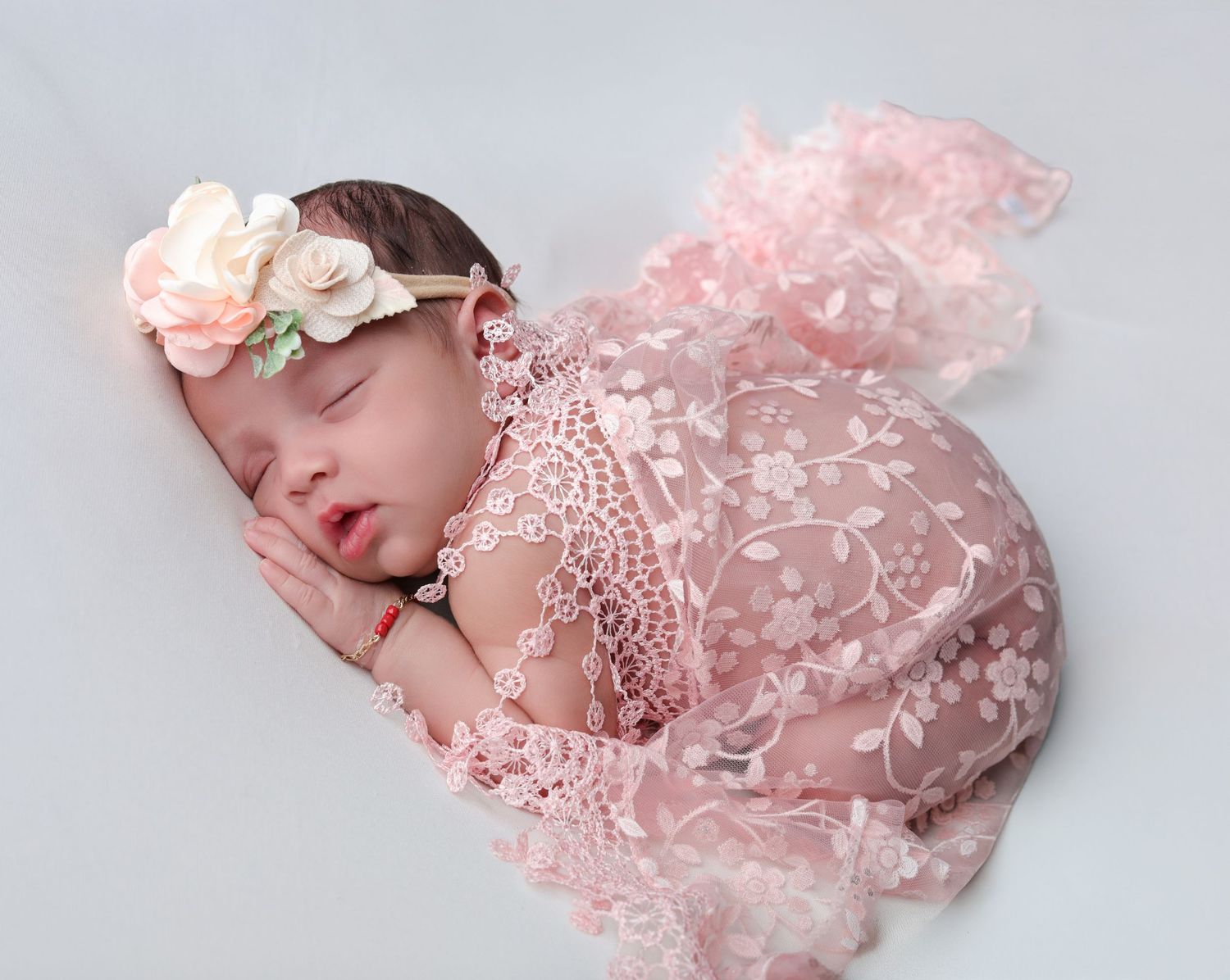 Sleeping newborn baby wearing a pink floral lace outfit and flower headband on a white background.