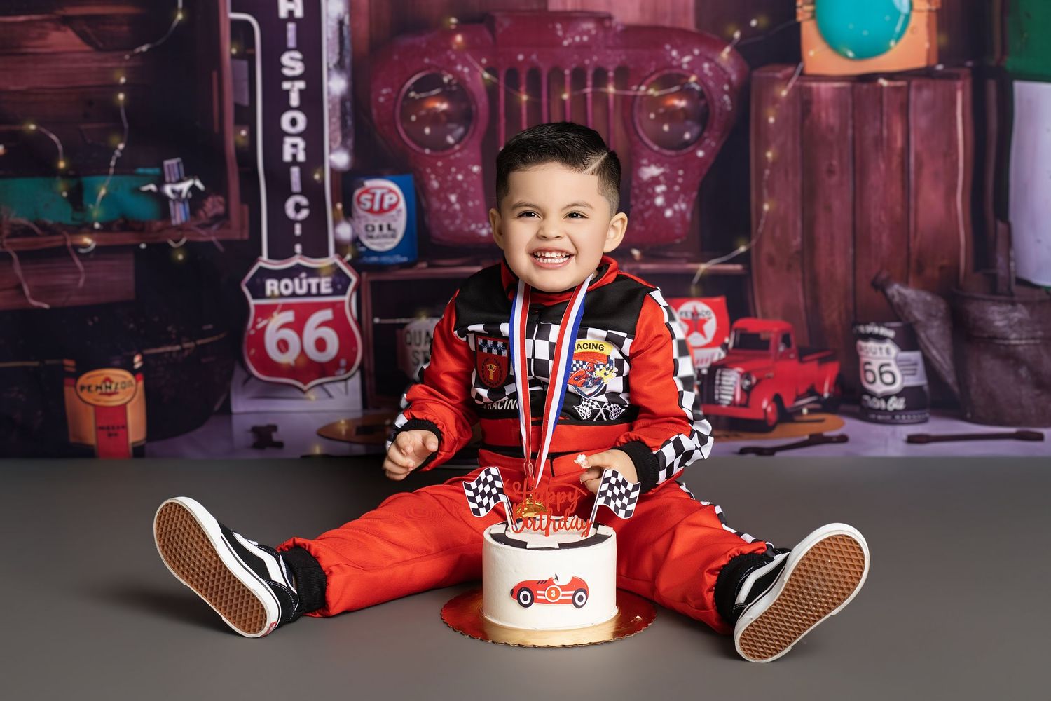 Smiling toddler in red racing outfit sits with birthday cake in front of Route 66 backdrop.