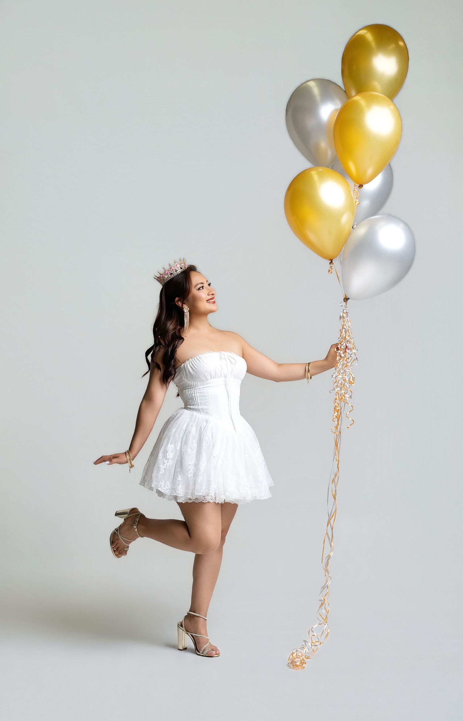 Young woman in white strapless dress and crown holds gold and silver balloons, posing joyfully in a studio.