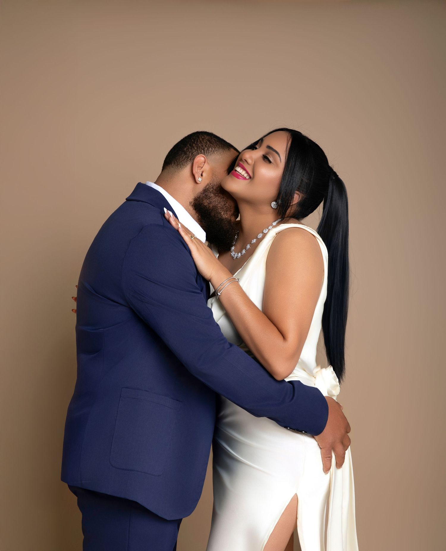 Couple in elegant attire embracing during engagement photo shoot, man in blue suit, woman in white dress with pearl necklace.