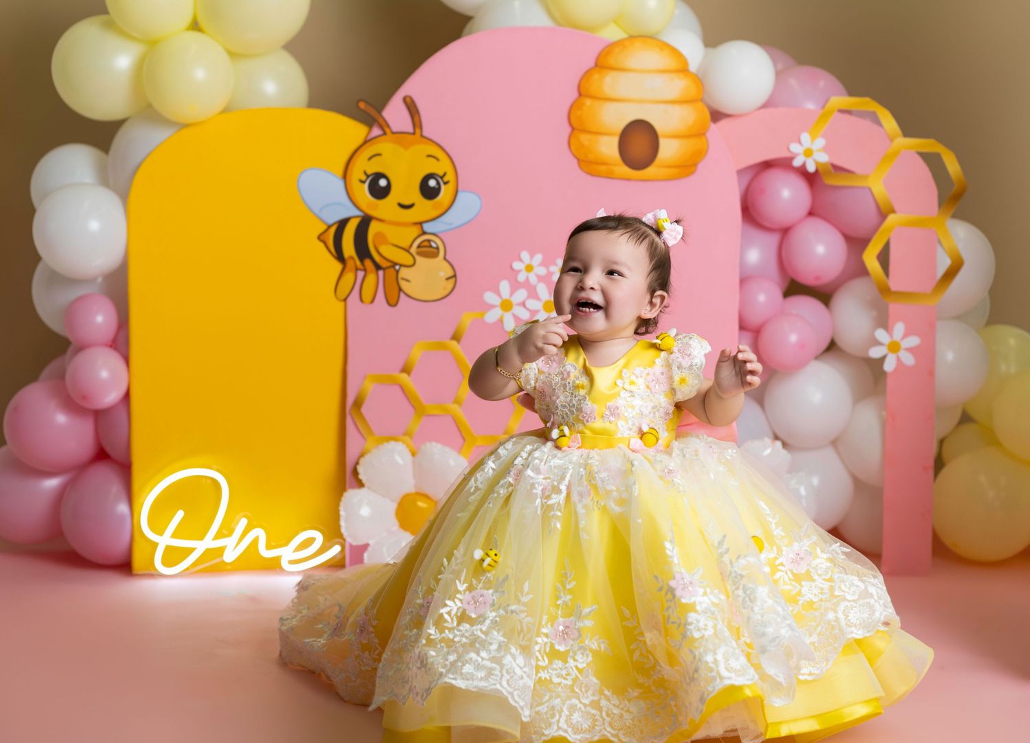 Baby girl in yellow floral dress celebrates first birthday with bee-themed backdrop, balloons, and honeycomb decorations.