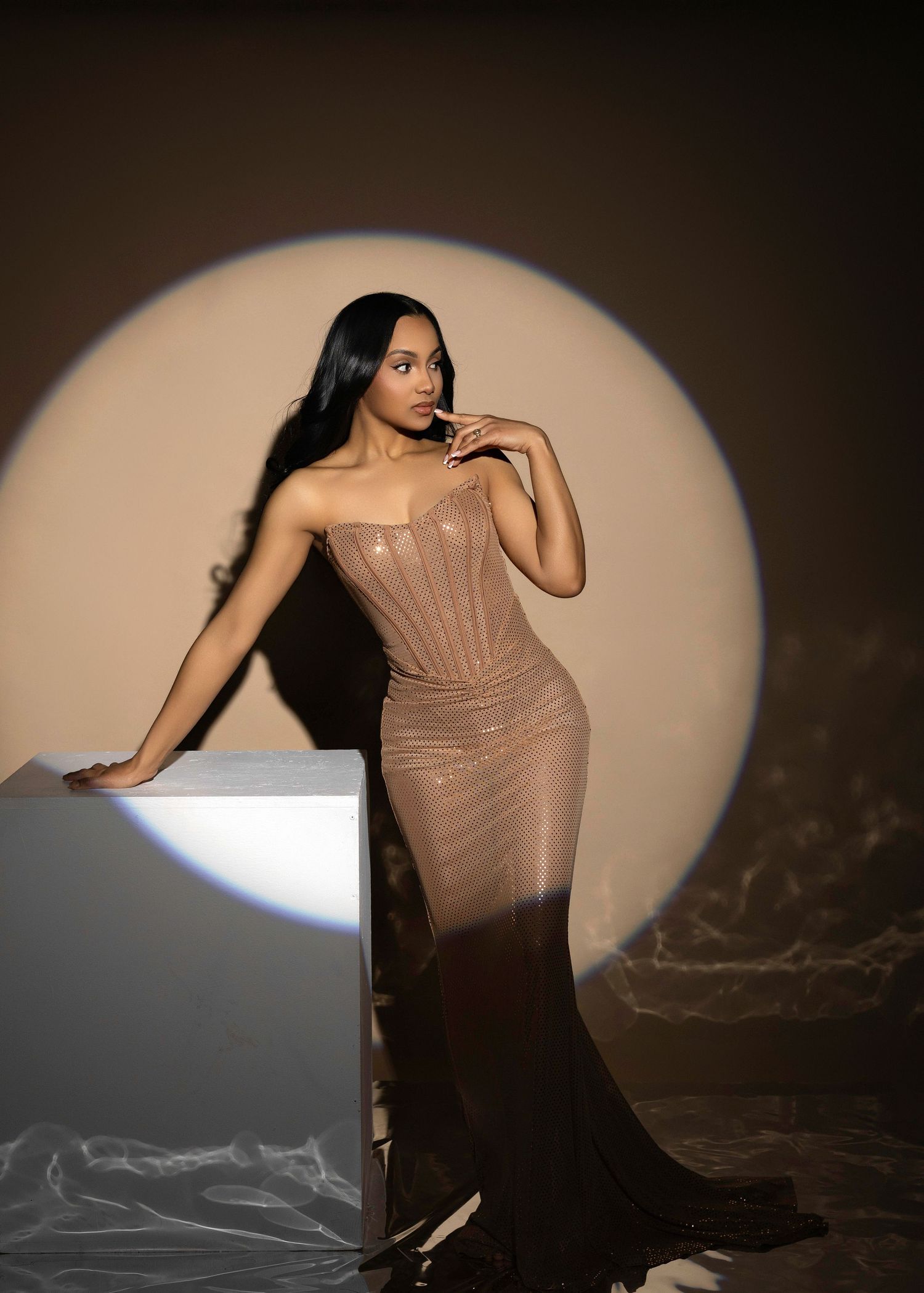 Woman in elegant brown corset gown poses gracefully against circular spotlight backdrop in dramatic studio setting.