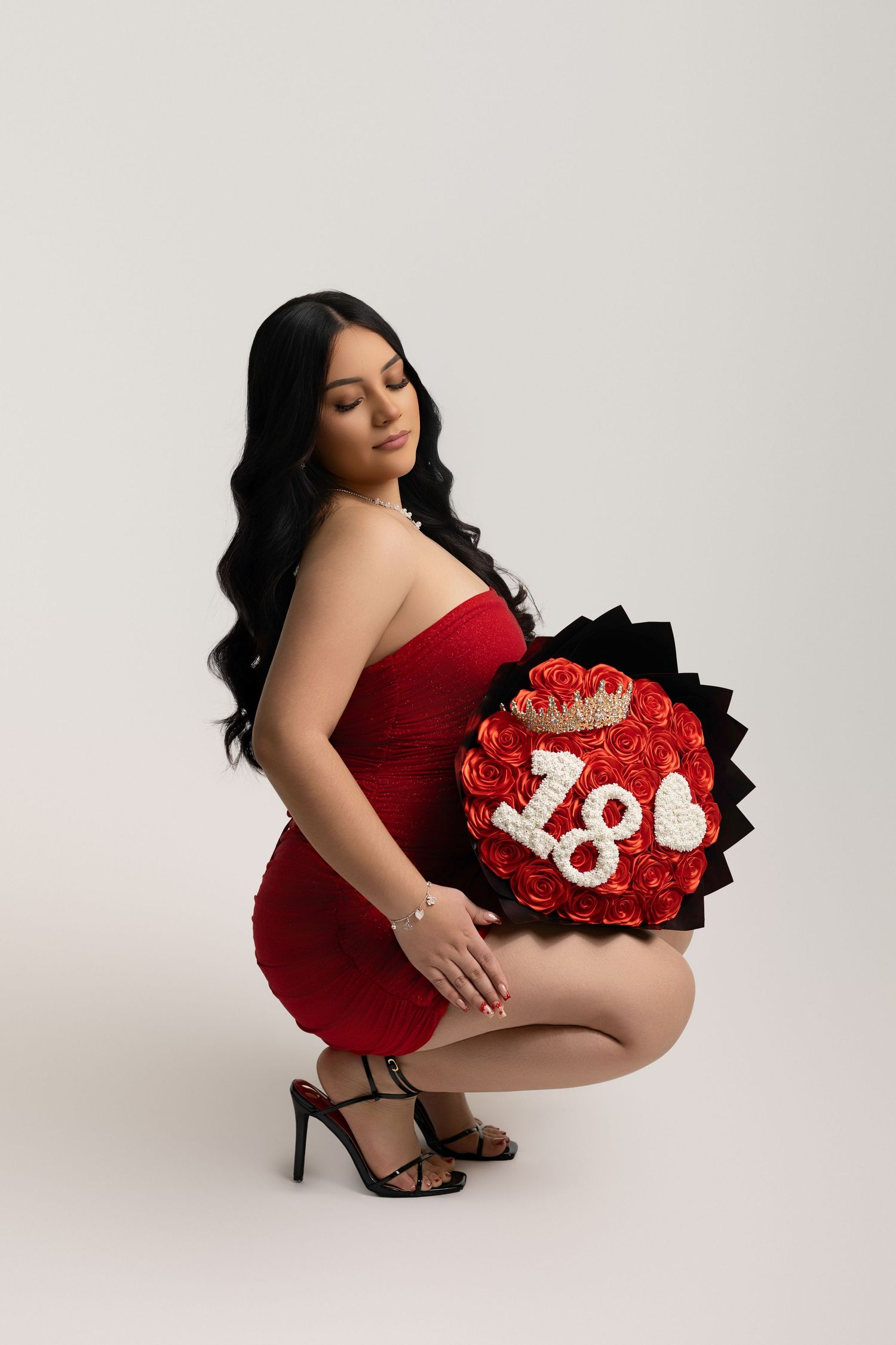 Young woman in red strapless dress crouching with a decorative rose bouquet displaying the number 18 and a crown.