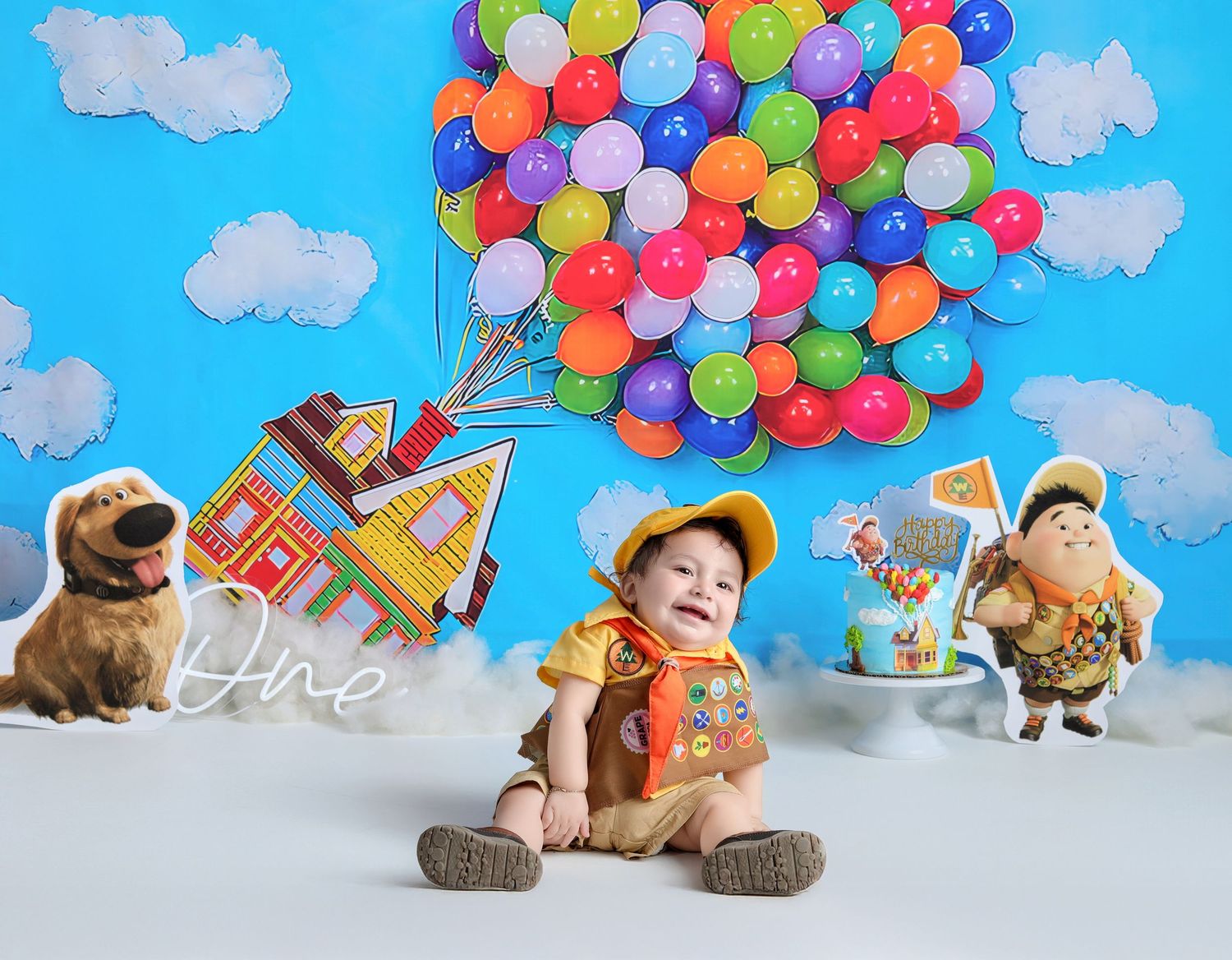 Baby dressed as Russell from Pixar Up sits before colorful balloon house backdrop with Dug dog cutout for first birthday.