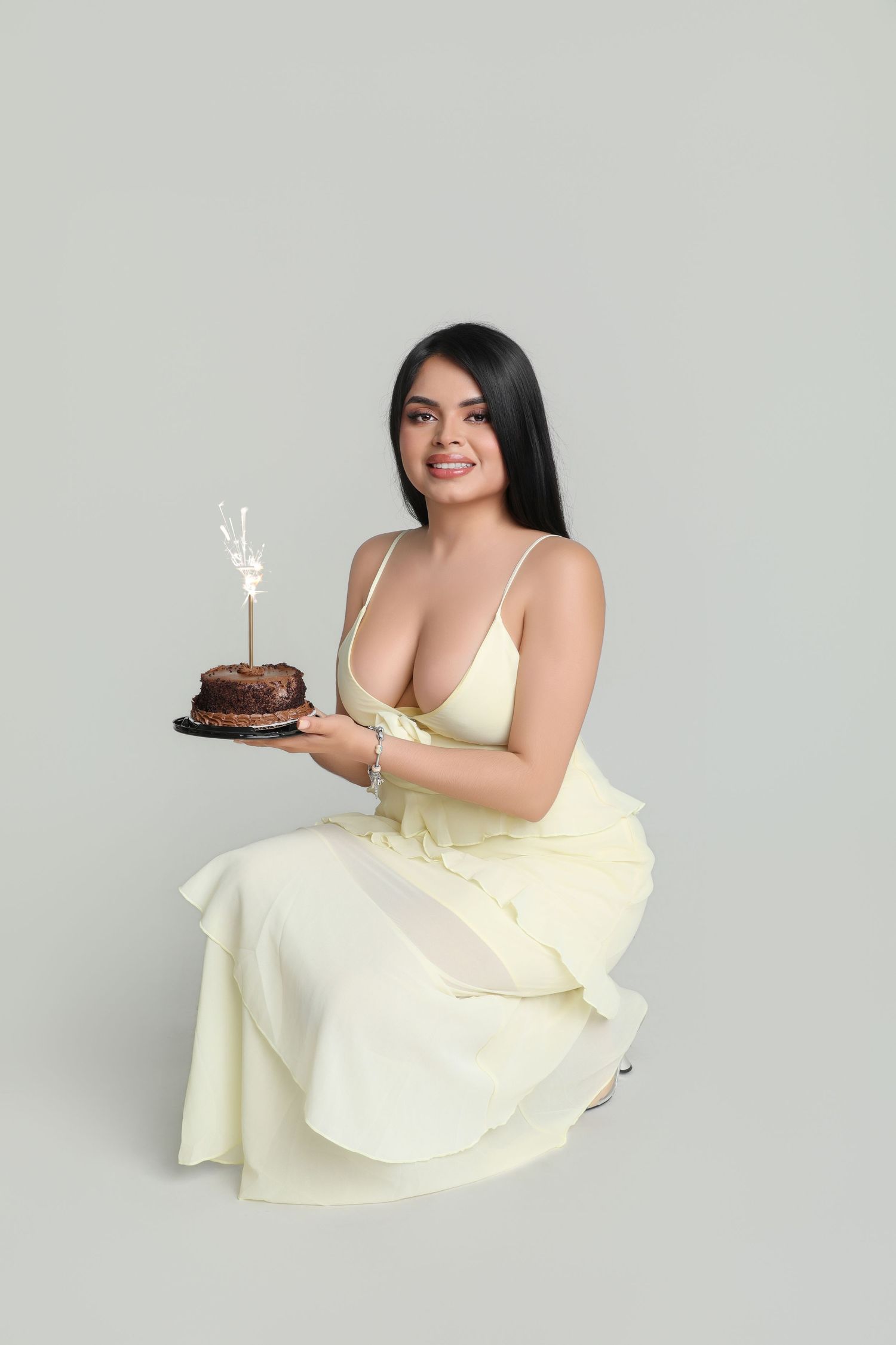 Young woman in cream slip dress holding chocolate birthday cake with sparkler candle, smiling at camera.