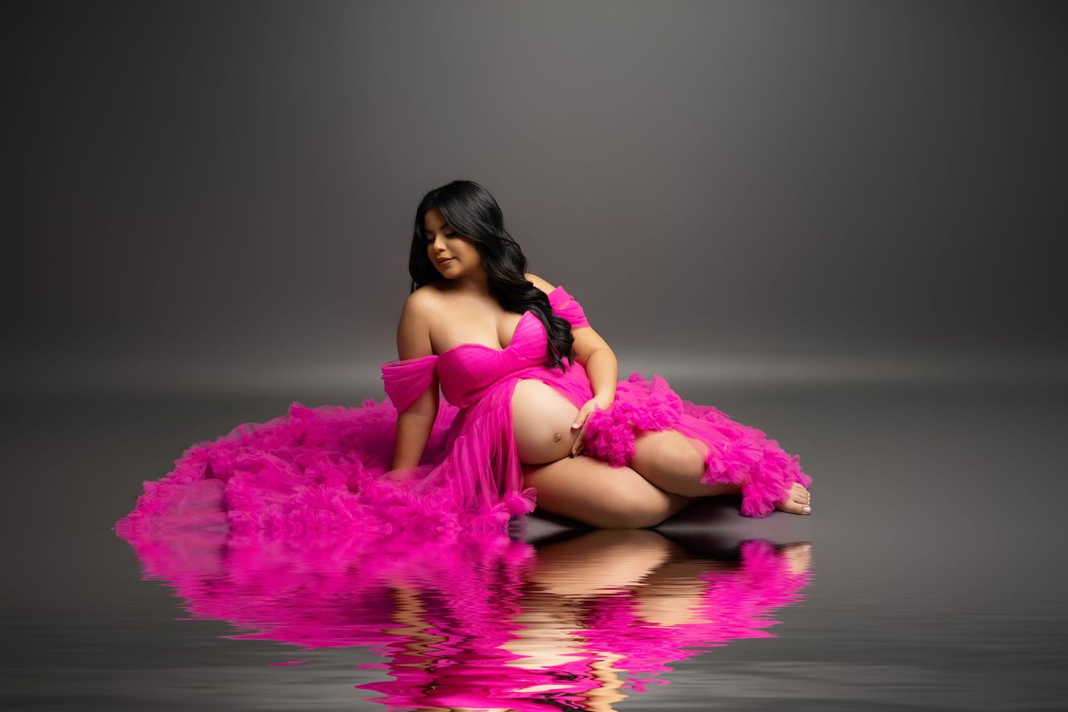 Pregnant woman in a flowing hot pink feathered gown poses gracefully on a reflective surface during a maternity photo shoot.