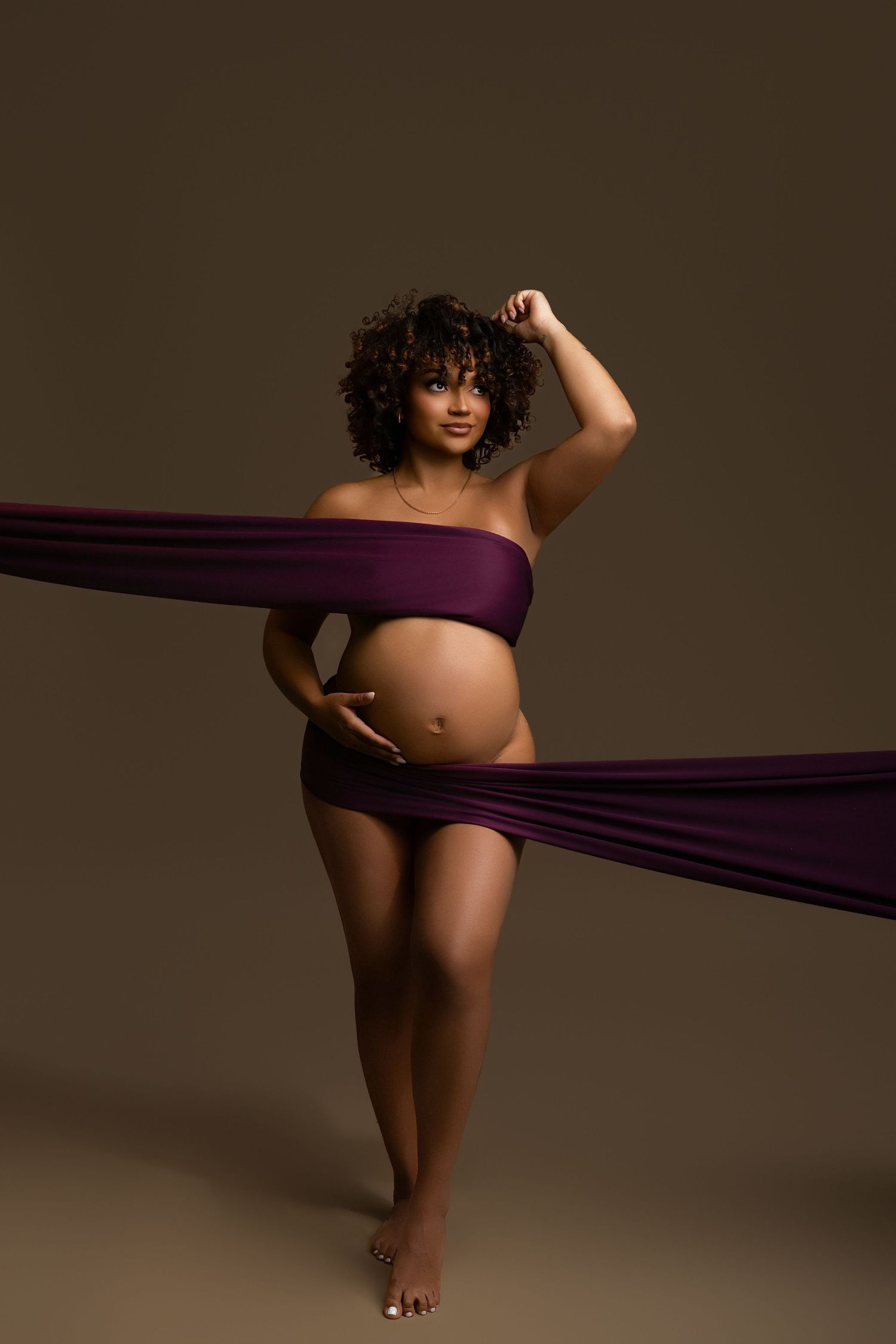 Pregnant woman wrapped in purple fabric, posing confidently in a studio with a brown background.