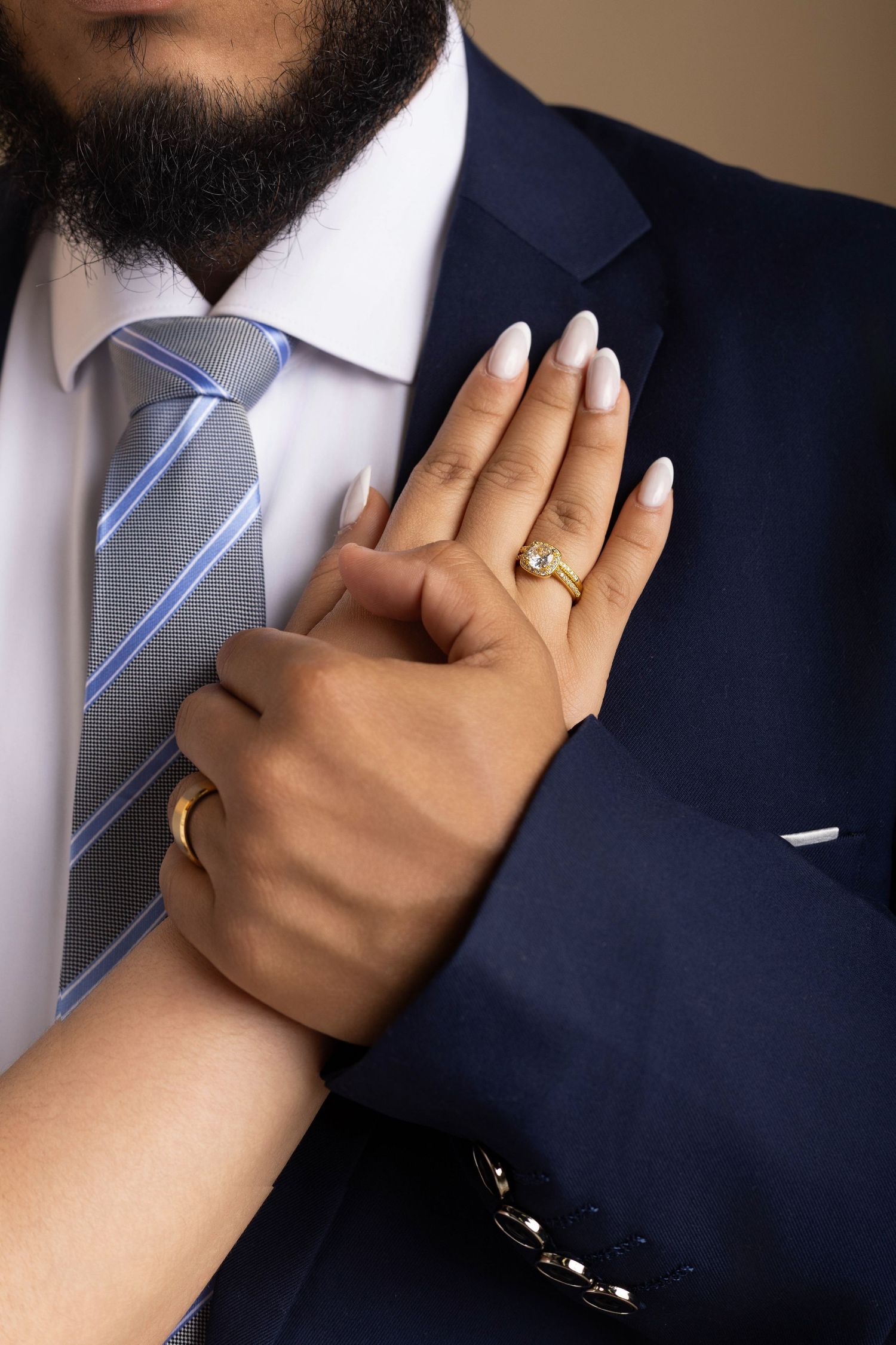 A woman's hand with gold rings and white nails rests on a bearded man's chest in a navy suit with striped tie.