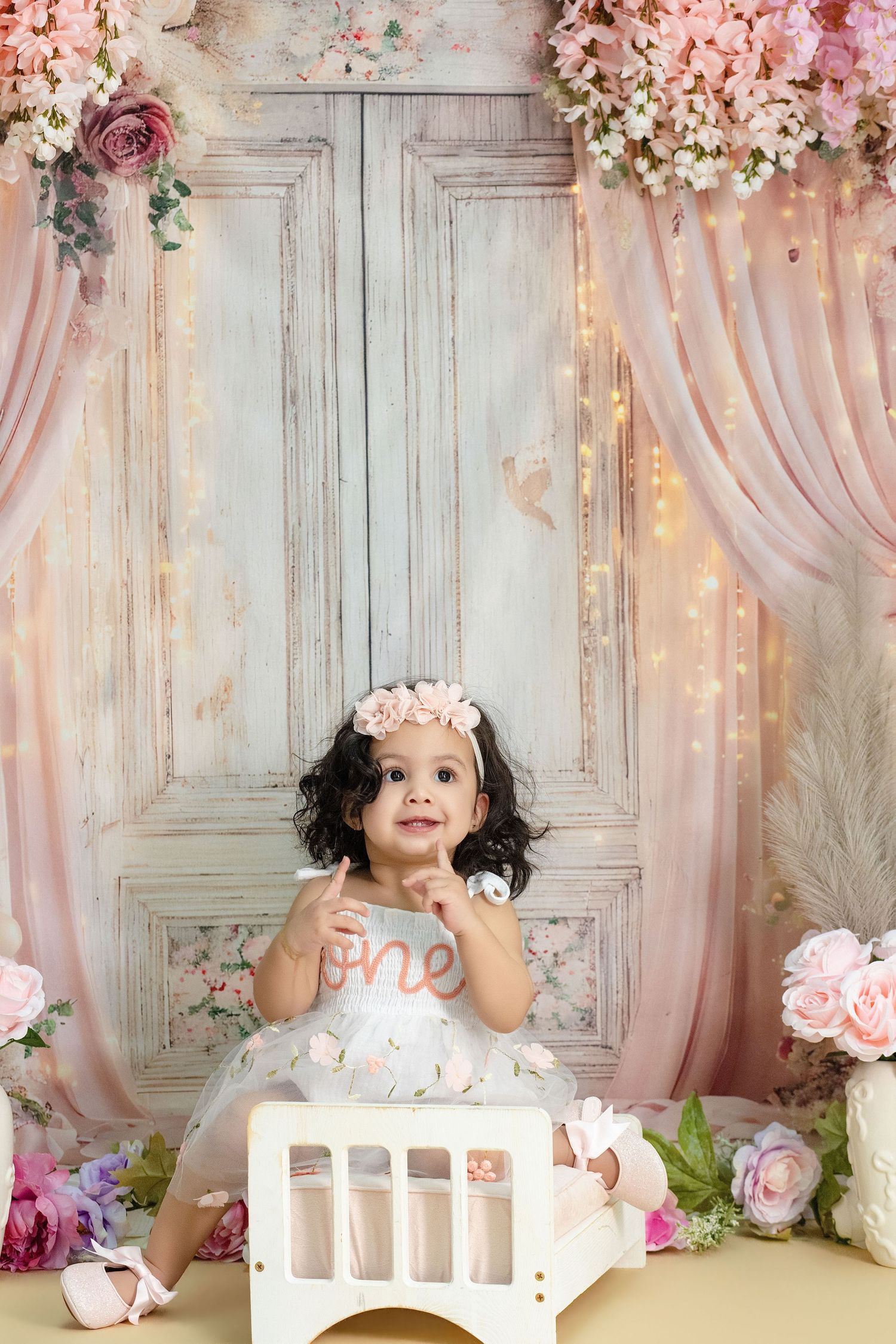 Adorable baby girl in white dress celebrates first birthday with floral backdrop and fairy lights.