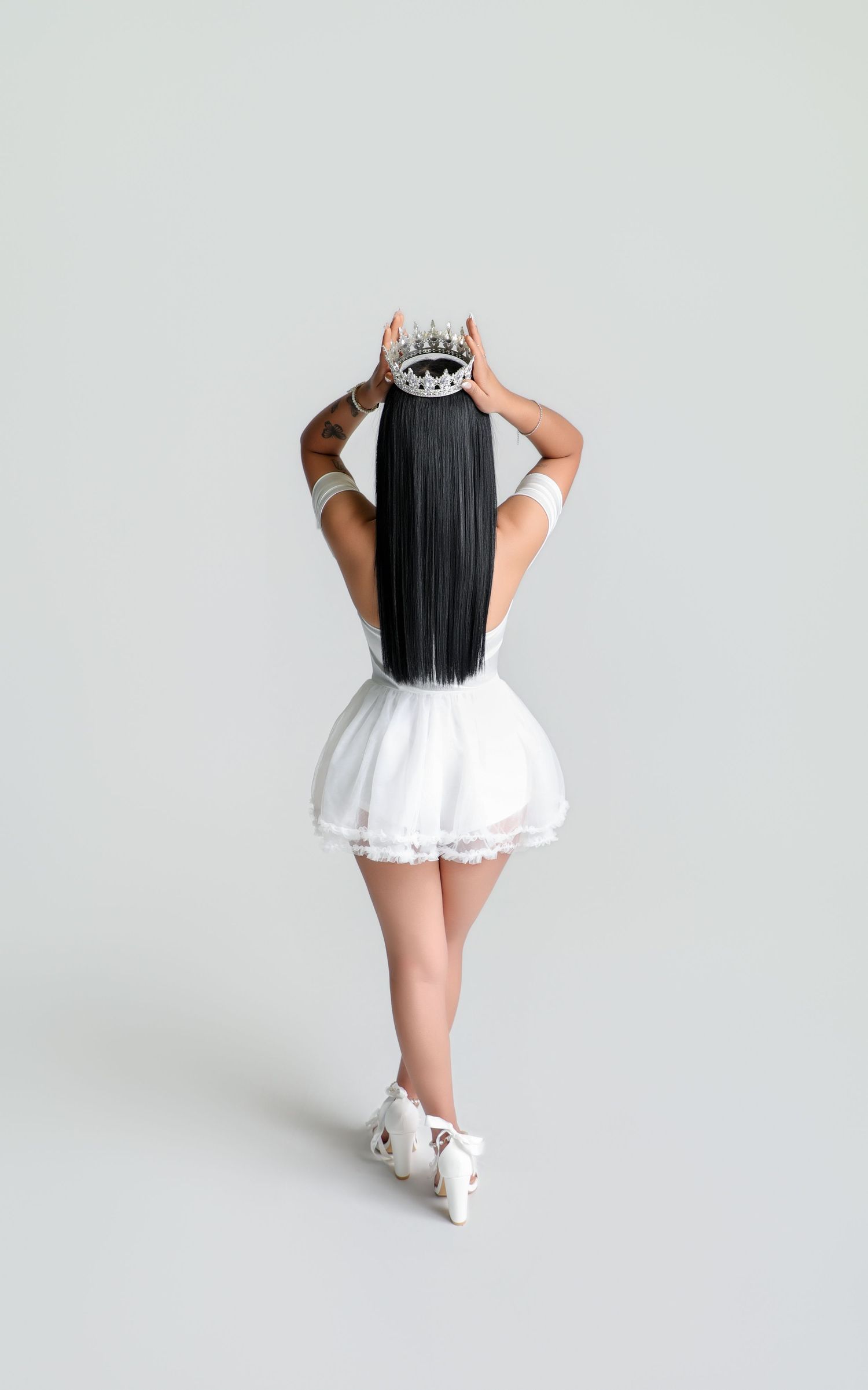 Woman in white mini dress holding a decorative crown above her long black hair, viewed from behind on gray background.