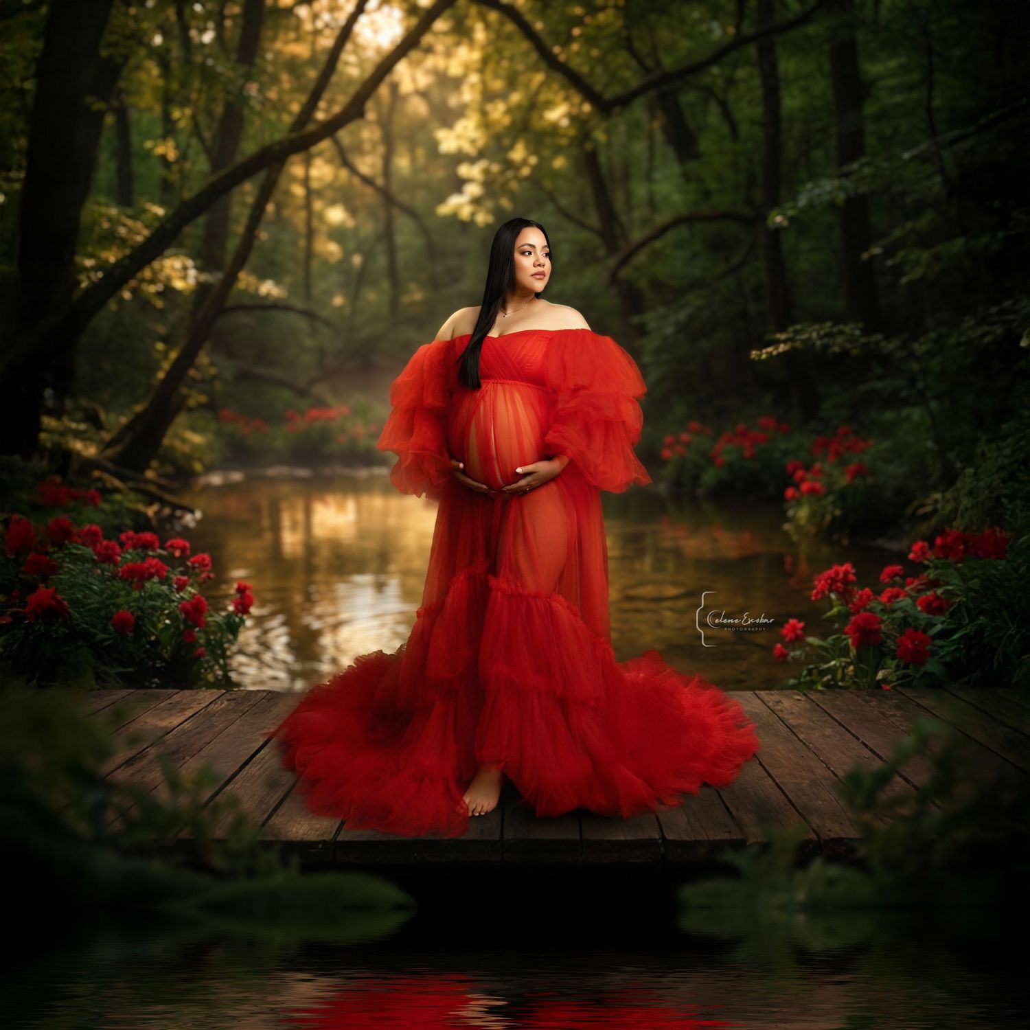 Pregnant woman in red tulle maternity gown posing on wooden dock surrounded by lush forest and red flowers.
