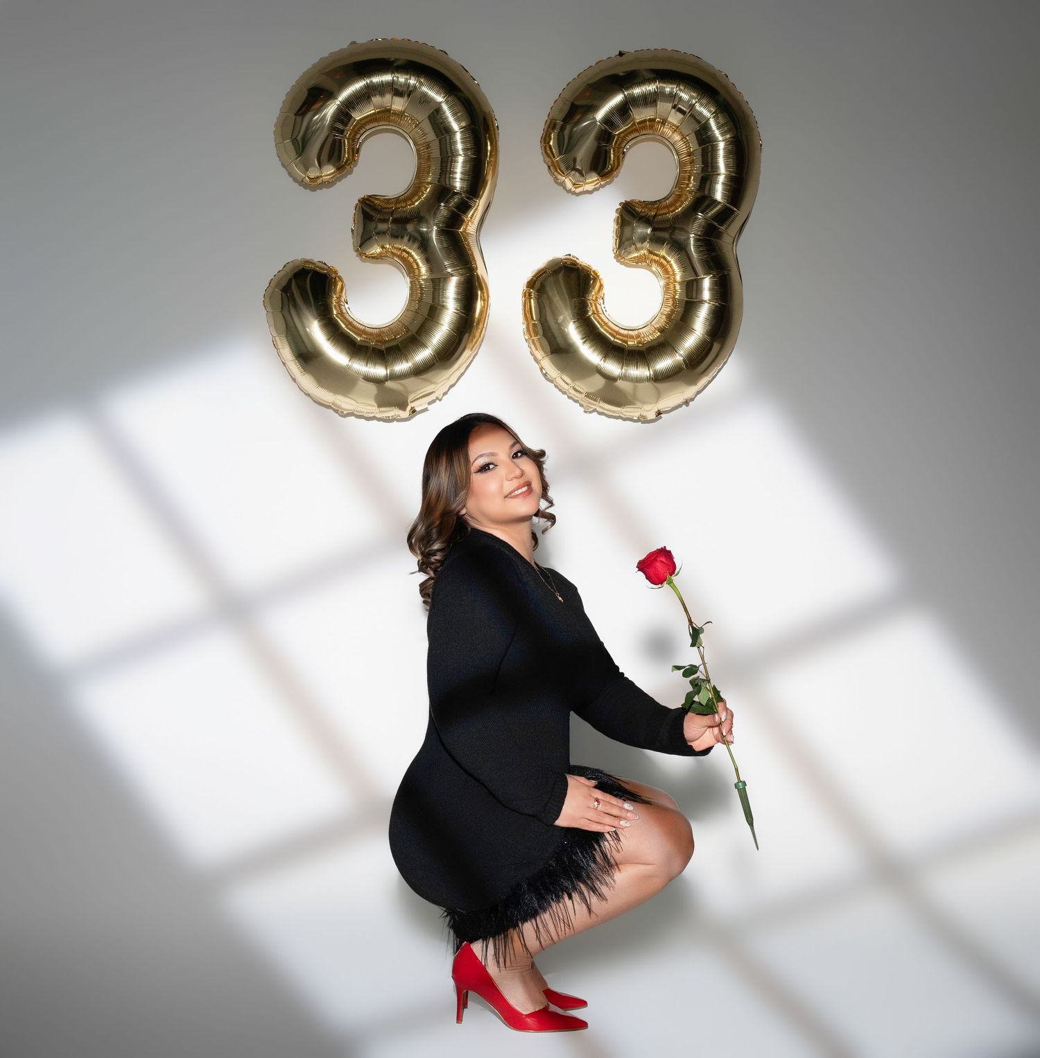 Woman in black dress holding a red rose poses with gold number 33 balloons celebrating her 33rd birthday.